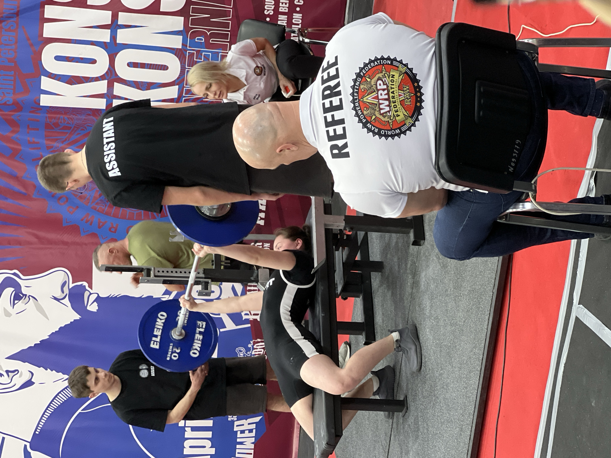 This image captures a powerlifting competition indoors in Saint Petersburg, Russia. The central focus is a female powerlifter, dressed in a black and white singlet, lying on a bench press attempting a lift. She is holding a barbell loaded with blue Eleiko brand weights, each marked "20" kg and "SWEDEN".
Surrounding her are several individuals in various roles. Directly behind her head, a male assistant in a black t-shirt with "ASSISTANT" printed on the back stands ready to spot the lift. Another male assistant in a black t-shirt is positioned near the barbell on the left, also seemingly ready to assist. Further back, another male individual in a green shirt is visible behind the bench press rack.
To the right, a bald male referee, wearing a white shirt with "REFEREE" prominently displayed on the back, is seated on a chair, observing the lift intently. His shirt also features a circular logo with text including "WORLD RAW POWERLIFTING", "WRP FEDERATION", "PER ASPERA AD ASTRA", and "RAW FEDERATION". Another person, a woman with blonde hair wearing a white shirt, is partially visible sitting behind the referee.
The background features a large blue and red banner. Visible text on the banner includes "Saint Petersburg", "KON" (part of a larger word), "RAW POWERLIFTING", "35", "April", and "INTERN" (likely "International"). Other partial texts like "SQU", "AN BEN", and "ATLON C" suggest various categories of the competition (squat, bench press, deadlift). The floor is a dark gray material, with a red strip marking part of the competition area. The scene conveys a focused and competitive atmosphere typical of a powerlifting event.