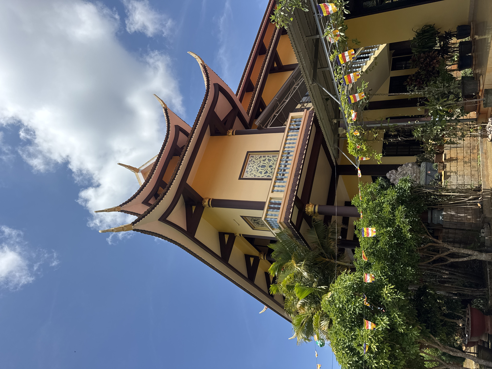 The image captures a low-angle, upward view of a temple in Lạc Dương, Vietnam. The temple features a traditional East Asian architectural style, characterized by a multi-tiered roof with curved eaves and decorative finials. The roof is primarily a tan color with a darker brown trim, and the structure below is a light yellow. A balcony with a blue railing is visible. Decorations resembling Buddhist prayer flags are strung along a cable or wire that is partially obscured by the foliage of the trees. There are potted plants and trees with green leaves and flowers. The sky is blue with scattered white clouds. The temple appears to be well-maintained, and the scene exudes a sense of cultural richness and serenity.