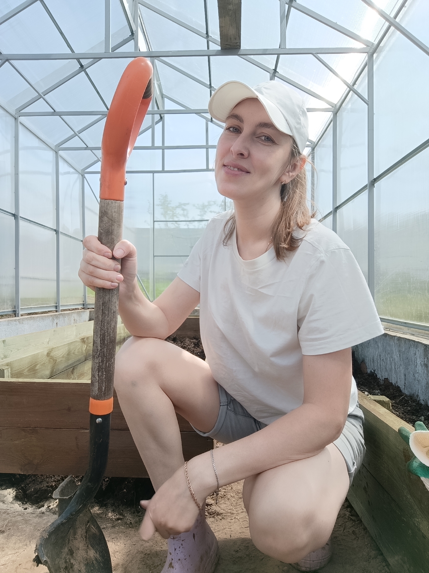 A woman with fair skin and light brown hair is squatting indoors, holding a shovel with an orange handle. She is wearing a white baseball cap, a white t-shirt, and gray shorts. A gold bracelet is visible on her left wrist. She is smiling slightly and looking towards the camera.
The woman is in what appears to be a greenhouse, with metal framing and translucent panels. Wooden boxes containing dark soil are present on the ground. The location may be Chaikovsky, Russia, but there is no information in the image to confirm this.