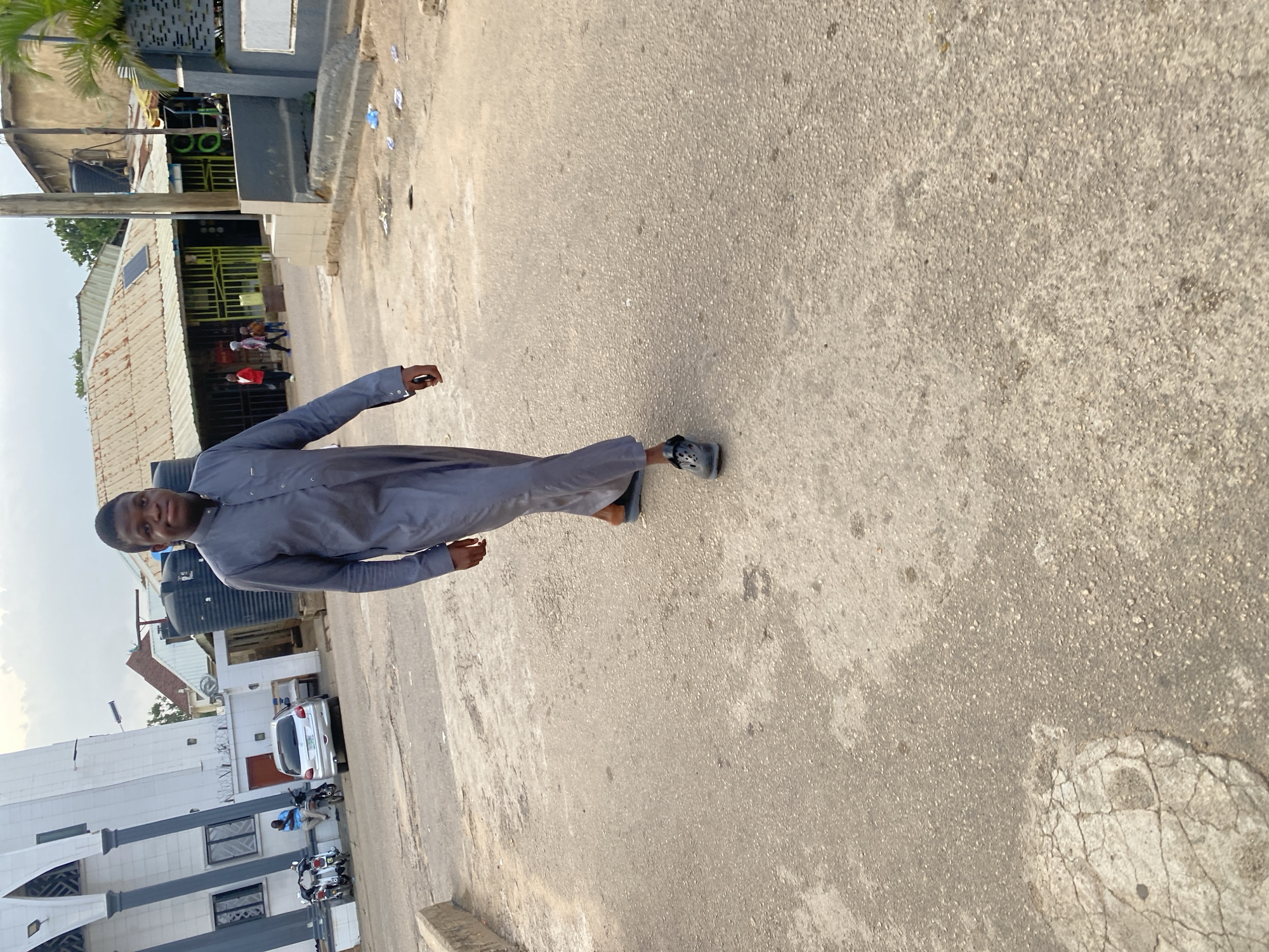 A young Black male is captured in a full body shot, walking towards the camera on an asphalt road. He is dressed in a gray traditional outfit, consisting of a long robe with a buttoned collar and loose sleeves. He is wearing dark, casual slip-on shoes. The man has a slight smile on his face.
The environment suggests a street scene in Jos, Nigeria. To the left of the frame, a white building with dark columns and arched windows, possibly a mosque, is visible. Several motorcycles are parked in front of it, and a person in a blue shirt is seated nearby. A silver sedan is parked further down the street. In the background, a row of buildings with corrugated iron roofs indicates a commercial or residential area. One building has solar panels installed on its roof. Water tanks are also visible, a common sight in Nigerian urban environments. The ground is rough asphalt with some cracks and debris, characteristic of city roads. The sky is overcast.
