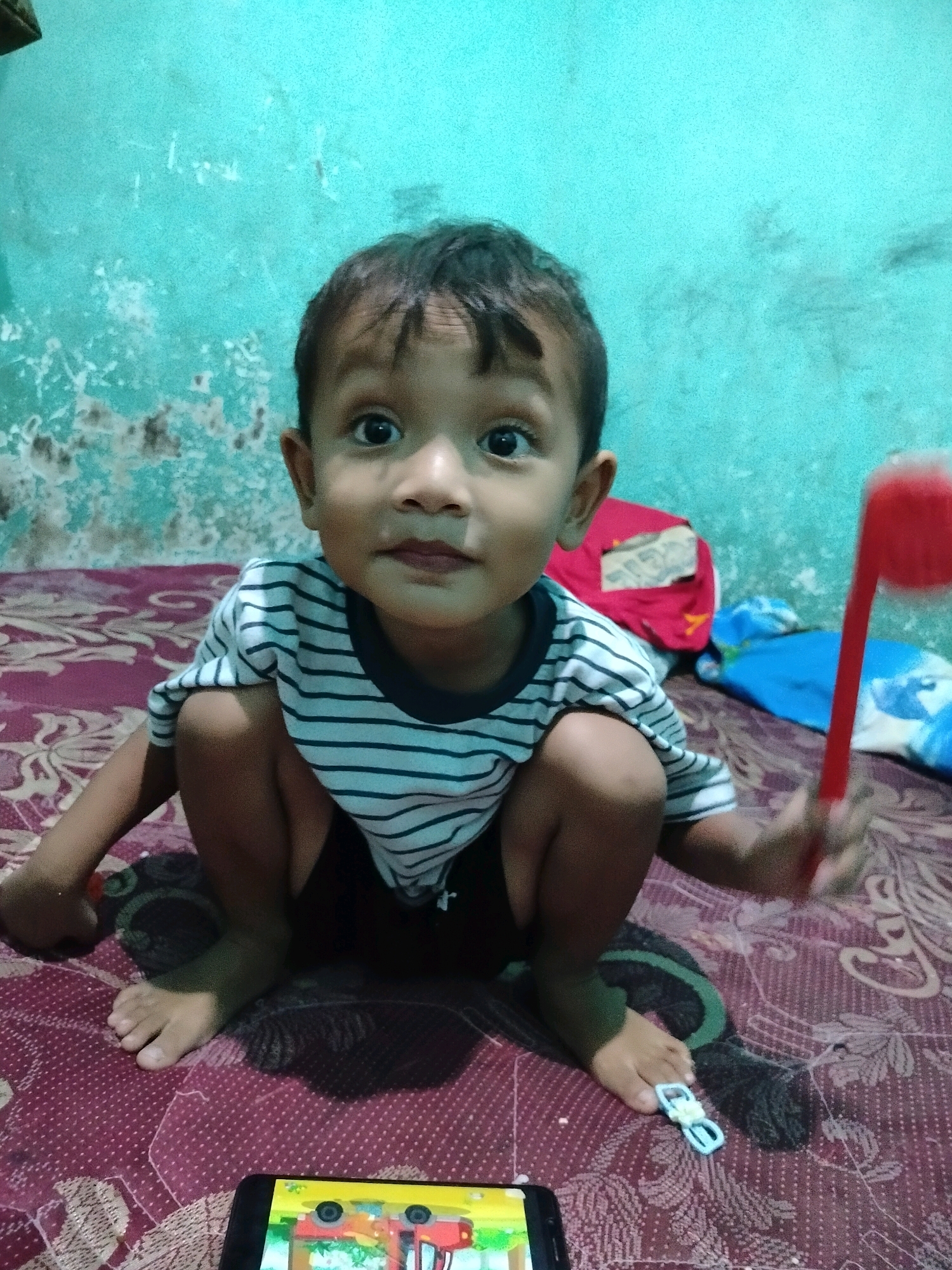 A young child with dark hair and dark eyes is crouched on a bed in Beloh, Indonesia. The child is wearing a black and white striped t-shirt and black shorts. The child is holding a red object in their right hand and is looking directly at the camera. The bed has a red and white patterned sheet. Behind the child, a turquoise wall with peeling paint is visible. A smartphone is placed on the bed in front of the child, displaying a colorful image.