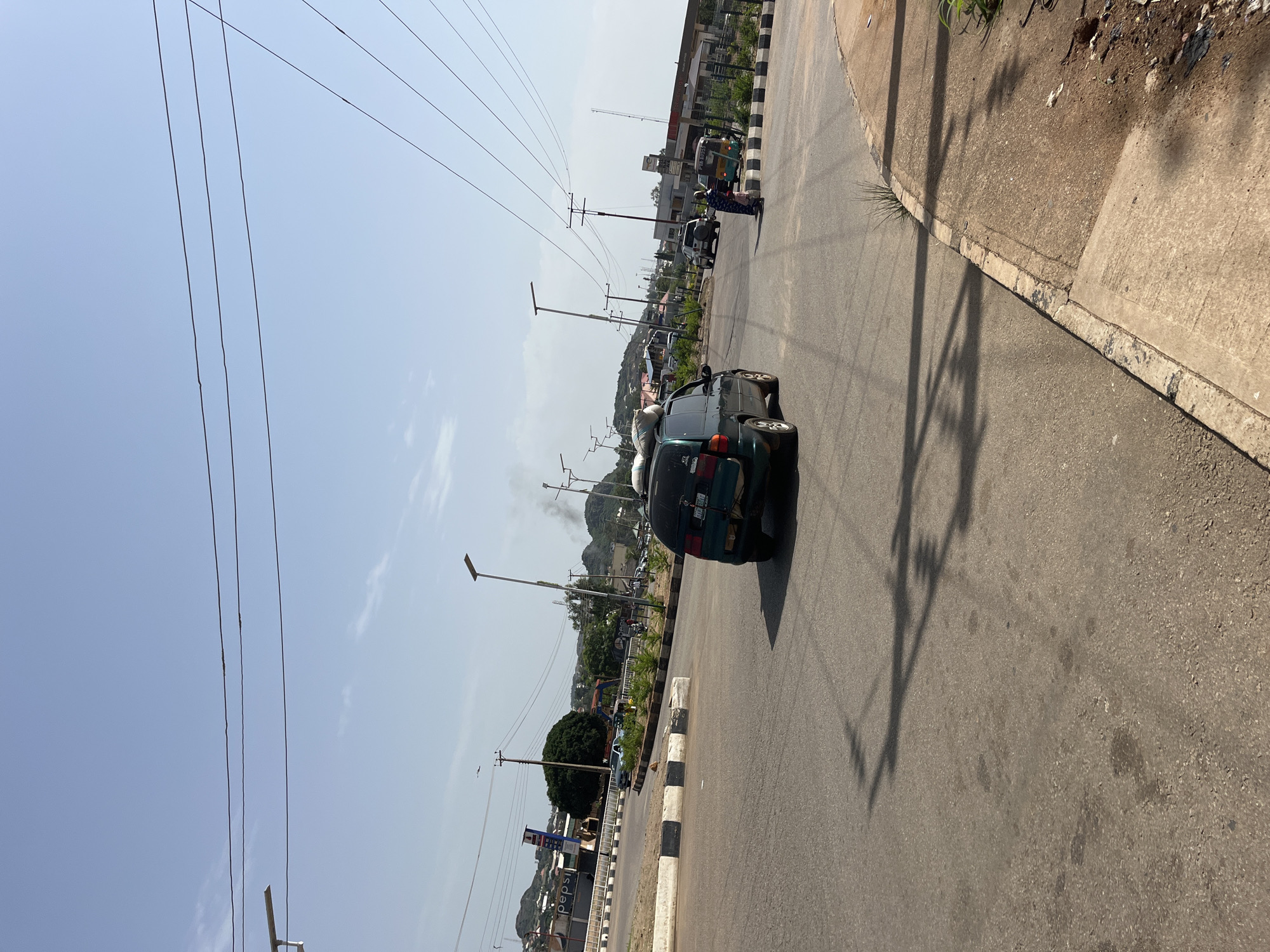 This media file captures a street scene in Bukuru, Nigeria, likely during daylight hours given the bright sun and clear sky. The image is oriented vertically, with the sky taking up a significant portion on the left and the road and surrounding environment on the right.
The foreground is dominated by a road with distinct black and white striped borders, suggesting a traffic calming measure or a marked lane. A dark green SUV is the most prominent object, parked partially on the road, with its rear visible. It appears to be carrying a load of bags on its roof. Further down the road, other vehicles are visible, including a tricycle and a white SUV. A few individuals can be seen near the vehicles. One person, possibly a traffic official in a blue uniform, stands near the tricycle.
The background shows a mix of commercial buildings, including a petrol station with signage that includes "PPC" and "PEPSI." There are also trees and hilly terrain in the distance. Overhead, a network of power lines stretches across the sky, supported by poles. The lighting suggests it's a sunny day, casting distinct shadows from the power lines onto the road. The overall atmosphere appears to be that of an active, everyday street scene in a Nigerian urban or semi-urban area. No specific event is clearly depicted, but the presence of vehicles and people indicates normal daily activity.
