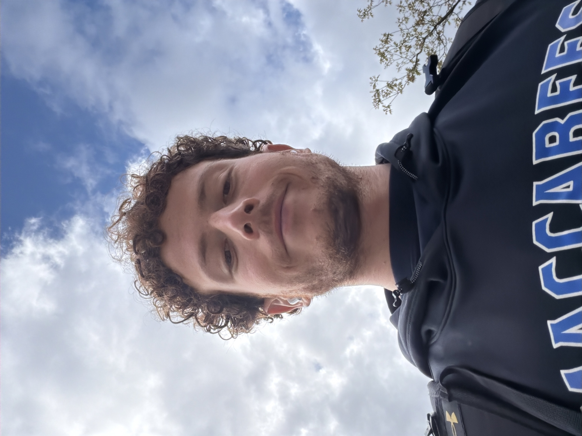 The image is a low-angle selfie featuring a young man with curly brown hair and a light beard, looking down towards the camera with a subtle, pleasant smile. He is wearing a dark-colored hooded sweatshirt with a blue and white design on the right side. Large block letters spelling "MACCABEE" are clearly visible vertically along the side of the sweatshirt. He is also wearing a dark backpack; a gold-colored logo resembling the Under Armour symbol is partially visible on a lower pocket of the backpack.
The setting is outdoors during the daytime, with a bright sky overhead. The sky is partly cloudy, displaying both patches of blue and fluffy white clouds. In the upper right corner of the frame, a few tree branches with light-colored leaves can be seen against the sky. The overall impression is a casual, relaxed moment, possibly capturing a quick outdoor snap. The man's expression conveys a sense of ease or contentment.