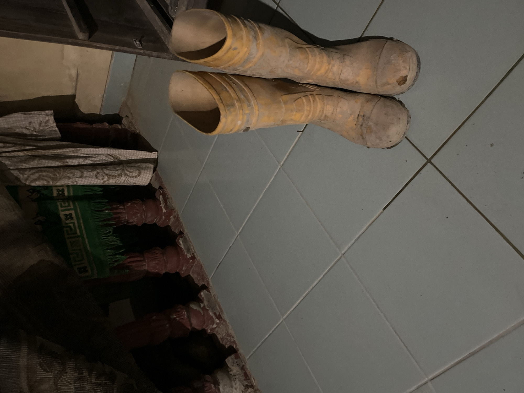 Two pairs of yellow rubber boots are placed on a tiled floor. The boots are worn and dirty, suggesting they have been used for work or outdoor activities. One pair is positioned slightly in front of the other, with the opening of the boots facing upwards. The floor is made of light blue tiles, arranged in a grid pattern. To the left of the boots, there are some folded fabrics and a green rug with a decorative pattern. The background is dark, with some wooden structures visible at the top. The lighting is dim, creating shadows and a somber atmosphere. The scene appears to be indoors, possibly in a storage area or a place where work boots are kept. There is no visible text in the image. The overall impression is one of practicality and utility, with the worn boots hinting at a life of labor or outdoor adventures. The location context is Kota Kuala Simpang, Indonesia, which might suggest a tropical climate where such boots would be useful for dealing with mud or wet conditions.