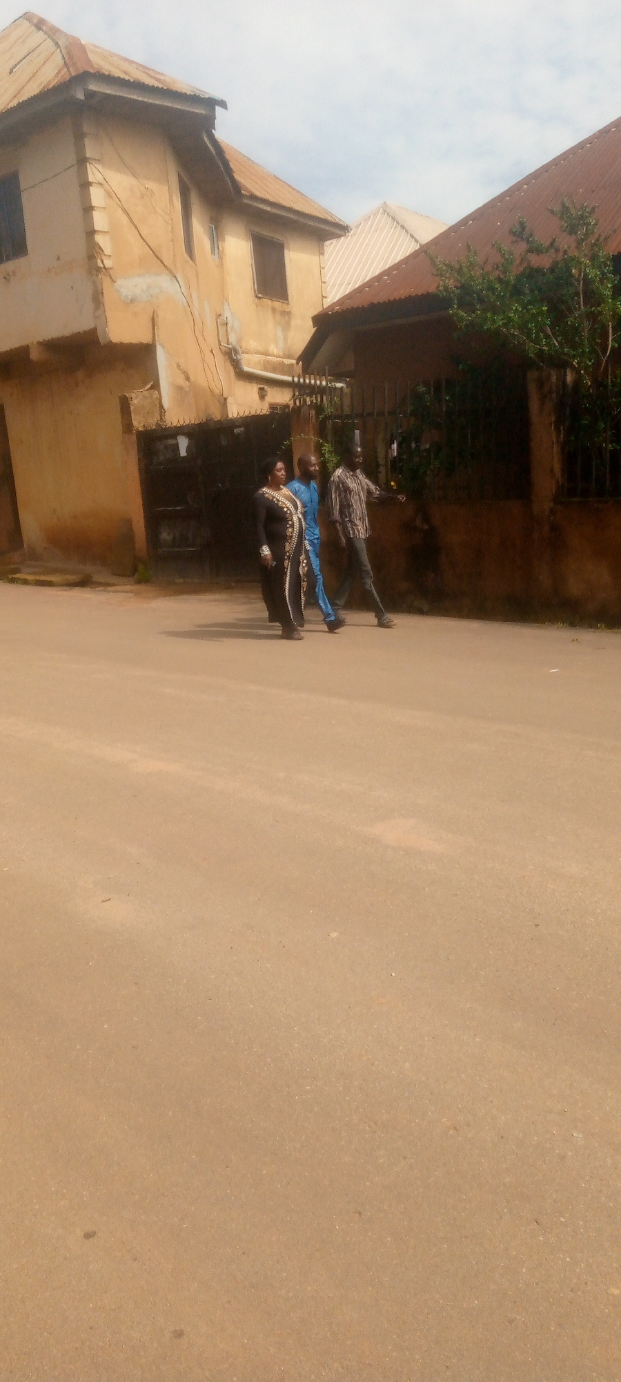 Three people are walking across a dirt road in front of a weathered, two-story building with a corrugated metal roof. The person on the left, a woman, is wearing a black and gold patterned dress. To her right, a man in a blue outfit walks behind her. Another man in a striped shirt and dark pants is to the right of the man in blue, his left arm extended forward as if gesturing. The building to their left appears to have a dark metal gate. The road is dusty and uneven, with sparse vegetation visible behind a dark metal fence on the right side of the image. The sky above is overcast.