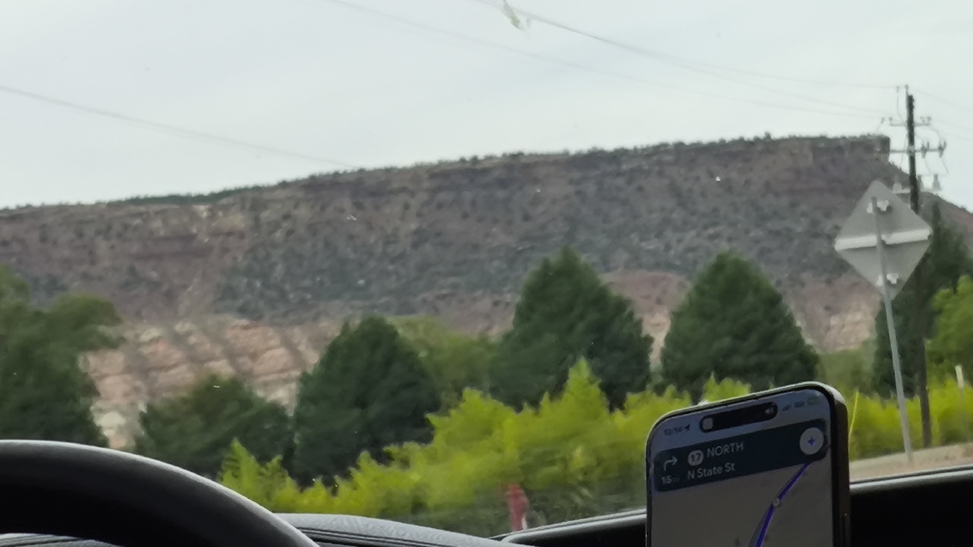 The image shows a view from inside a vehicle, looking out through the windshield at a mountainous landscape under an overcast sky. In the foreground, the steering wheel of the car is visible at the bottom left. A smartphone is mounted on the dashboard on the right, displaying a navigation app. The app shows the route heading north on N State St, with a distance of 15 miles remaining. A diamond-shaped road sign with a white cross shape is visible to the right of the phone, positioned in front of a utility pole. The middle ground is filled with lush green trees of various sizes, including tall, conical evergreens and lower, bushier foliage. Behind the trees, a massive, layered rock formation stretches across the horizon, exhibiting shades of brown, tan, and red, with visible striations and vegetation sparsely dotting its surface. The overall scene suggests a scenic drive through a natural area, likely in or near Rockville, United States of America, given the information on the navigation app.