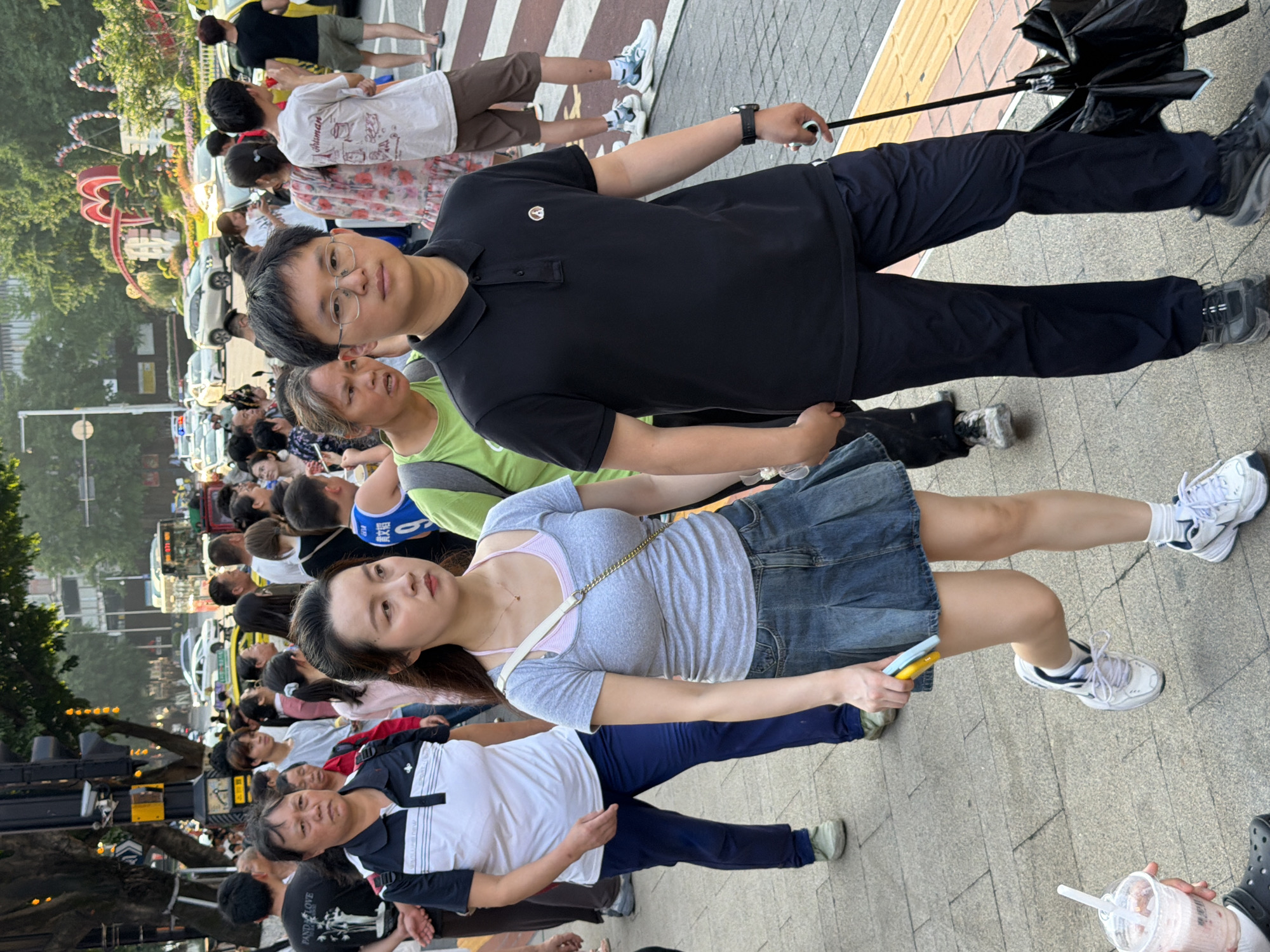 In Yuzhong District, China, a man wearing glasses and a black polo shirt walks with an umbrella in his right hand. Beside him, a woman in a grey t-shirt and a denim skirt walks while holding a yellow and blue object. Other pedestrians are visible in the background on a sidewalk, with traffic and buildings lining the street.