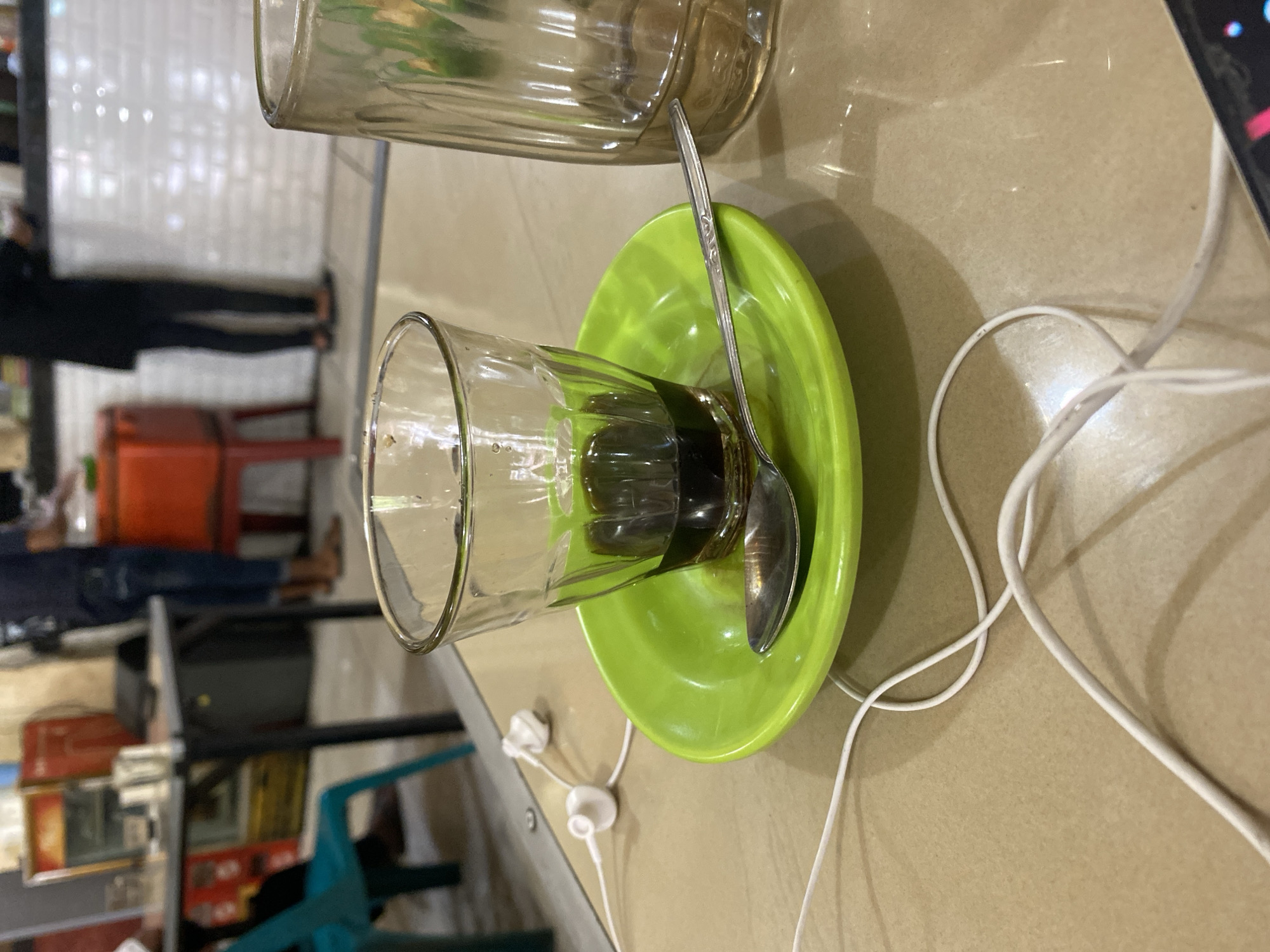 A close-up, eye-level shot captures a small glass of dark liquid, likely coffee, resting on a bright green saucer. A silver spoon lies on the saucer, partially submerged in the liquid. To the right, a tall, clear glass with ribbed sides is visible. White earbuds with their cord are strewn across the tan-colored table in the foreground. The background is blurred, showing a seating area with chairs and tables, and people standing behind a counter. This image was taken in Lhokseumawe, Indonesia.