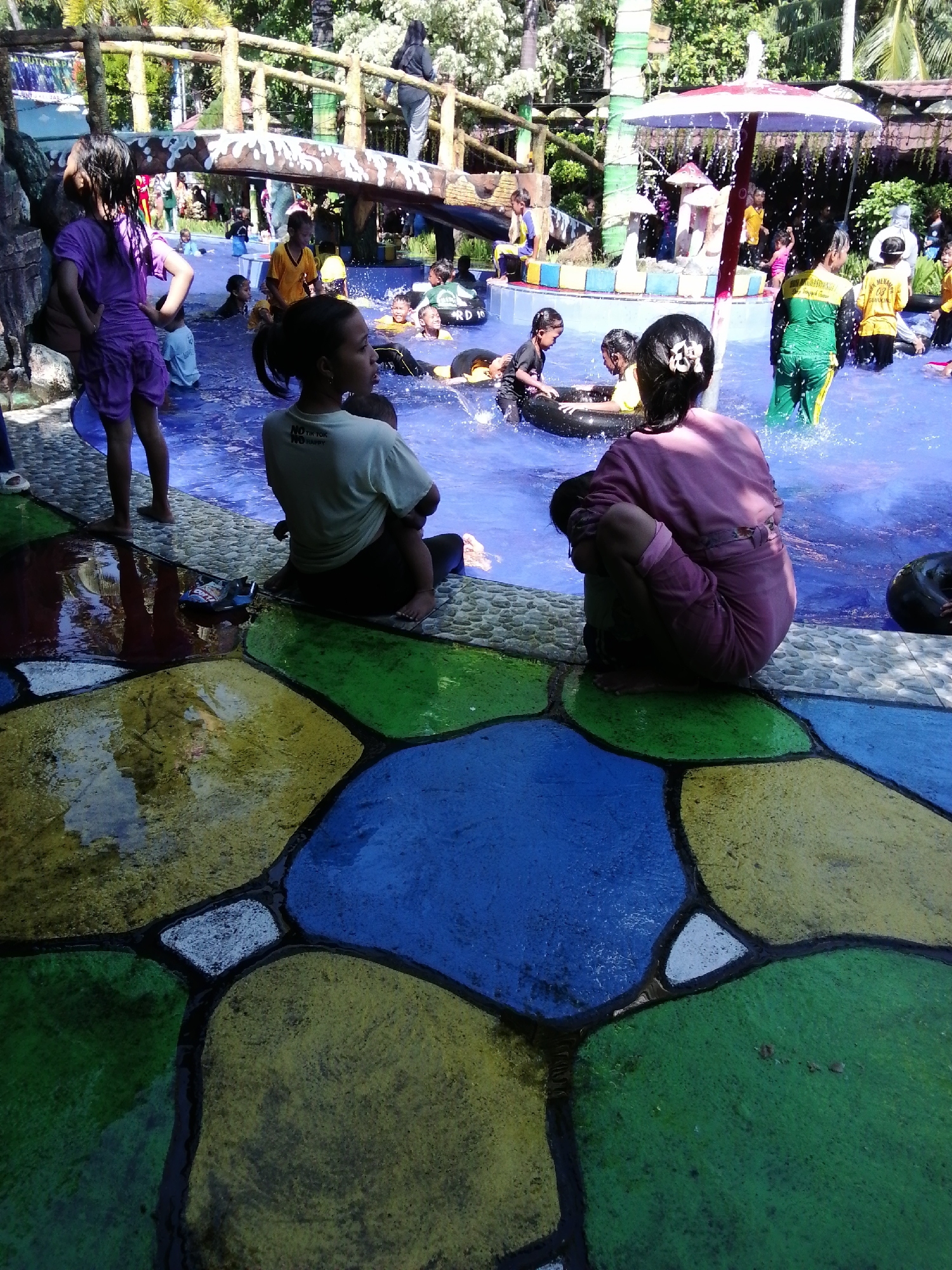 This is an outdoor, ground-level, medium shot of a water park in Tegalwangi, Indonesia. In the foreground, two women and a child sit on the colorful, wet concrete poolside. The woman on the left wears a light-colored t-shirt and holds a child in her lap. The woman on the right wears a pink tracksuit and hugs a child, who is curled up. Behind them, a group of children plays in the water. Some children sit on inner tubes, while others splash around. In the background, there is a wooden bridge with people crossing, and trees provide shade. The water is blue, and the pool deck is decorated with large, colorful shapes.