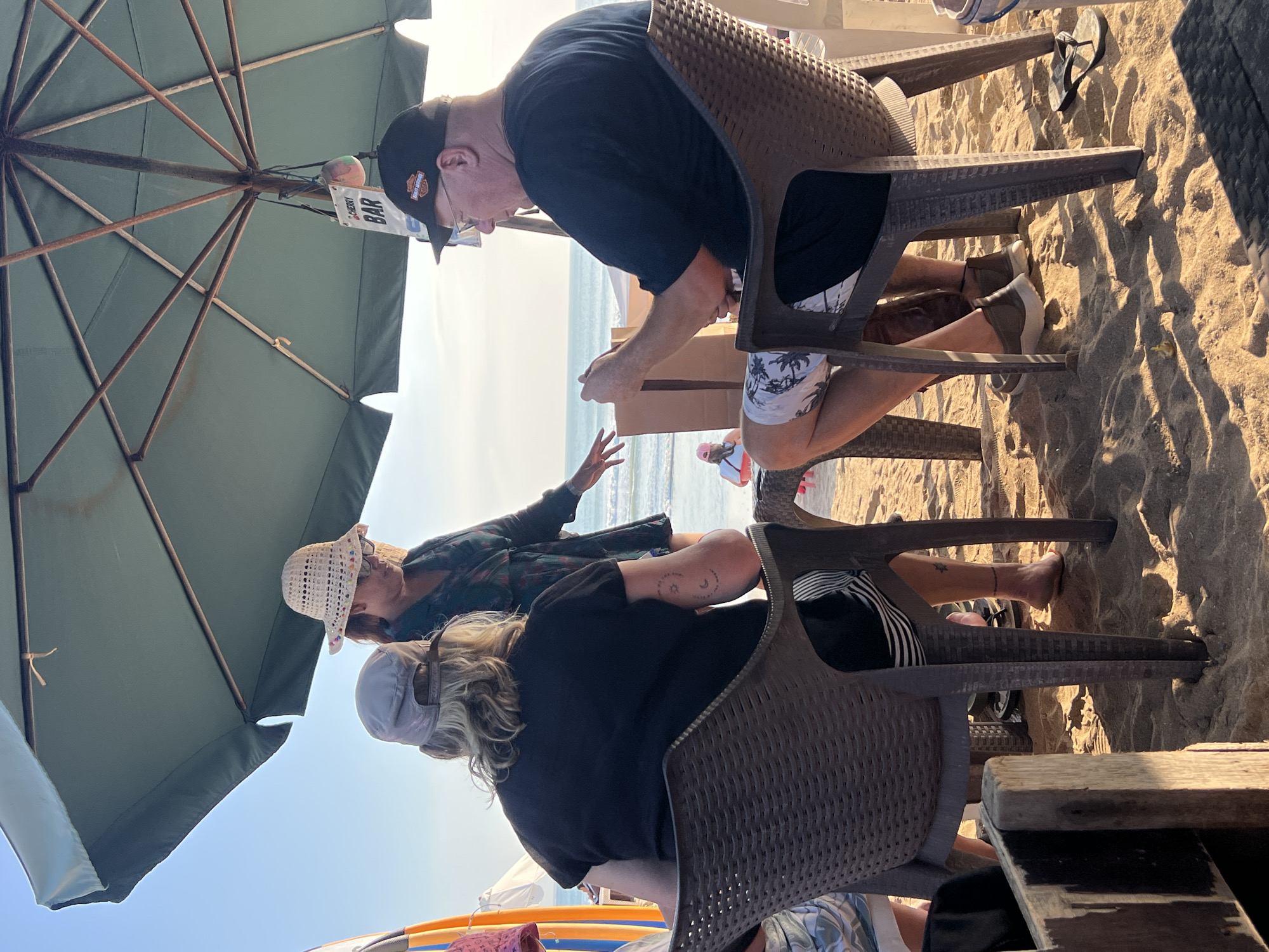 Three people are seated at tables on a sandy beach in Kuta, Indonesia, under a large, green umbrella. The person on the left, facing away from the camera, wears a black t-shirt and a gray baseball cap. The person in the center, facing the camera, wears a patterned dress and a straw hat. The person on the right, facing left, wears a black t-shirt, a black baseball cap with an orange logo, and patterned shorts. They are holding a mobile phone. The ocean is visible in the background with gentle waves. A sign hanging from the umbrella reads "BAR". Beach chairs and tables are set up on the sand, with some surfboards visible to the left of the frame. The sun is setting, casting long shadows across the sand.