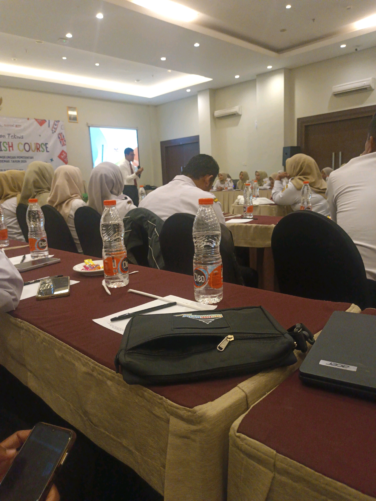 The image depicts an indoor event, likely a seminar or workshop, taking place in a room with a projector screen and ceiling lights. A male presenter, wearing a white shirt and dark pants, is seen speaking at the front of the room. A group of attendees, many wearing headscarves, are seated at tables in front of him. Several bottles of water, a smartphone, and a laptop are visible on the tables. The overall atmosphere appears to be formal and educational. The location is identified as Jogoloyo, Indonesia.