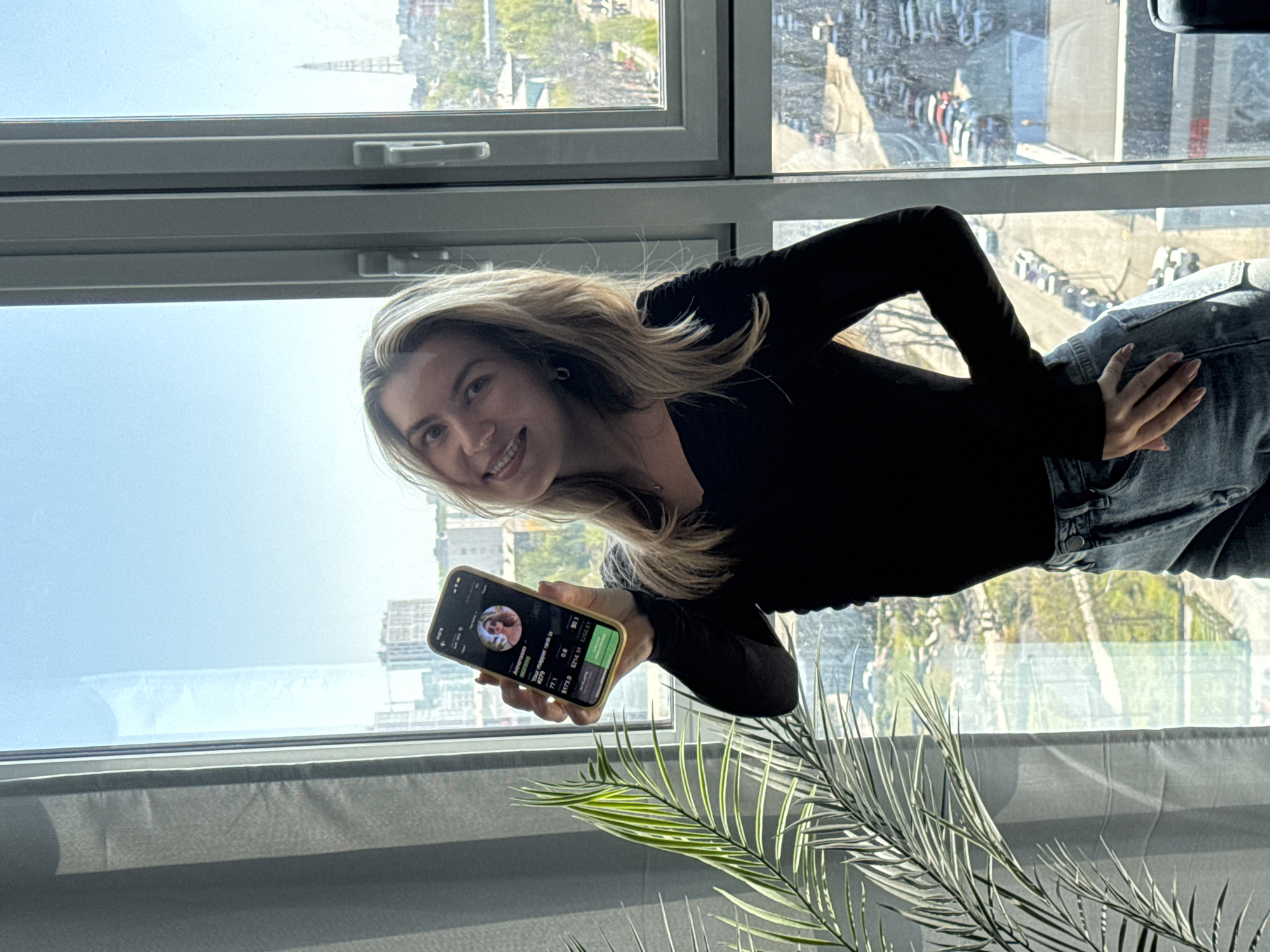 A woman stands in front of a large window, holding a cell phone displaying financial data. She is wearing a black long-sleeved shirt and blue jeans. The window offers a view of a city skyline, with buildings and vehicles visible. A potted plant sits on the windowsill.