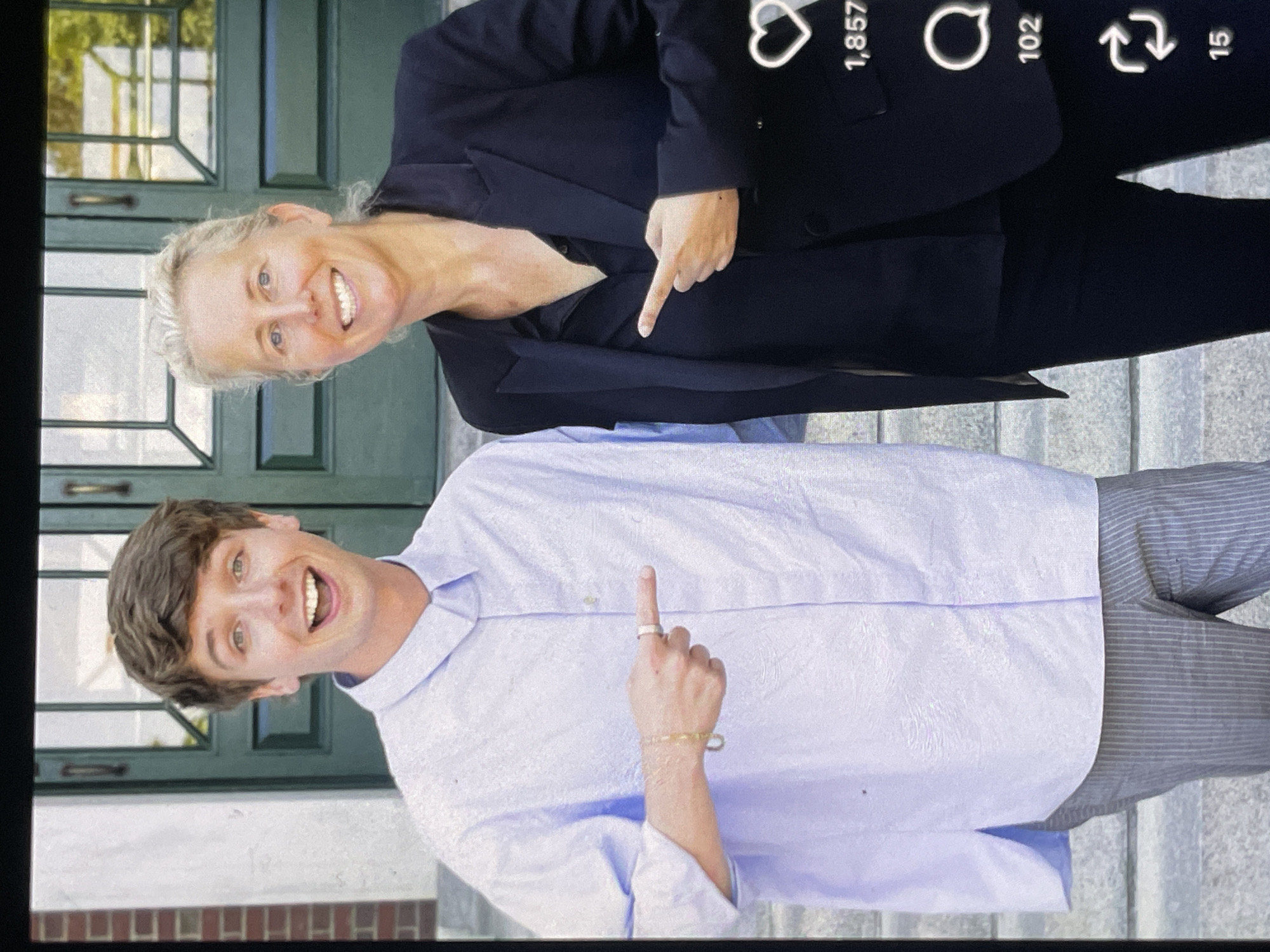 Two individuals, a young man with light brown hair and a woman with blonde hair, stand side-by-side in front of a dark green door with glass panels and a brick wall to the left. The man is wearing a light blue textured button-down shirt and grey pinstriped pants. He is smiling broadly and pointing his right index finger outwards. The woman is wearing a black blazer over a black collared shirt and black pants. She is also smiling and pointing her left index finger outwards, towards the man. They appear to be outdoors, possibly on a campus or in front of a building. There are icons and numbers visible on the right side of the image, suggesting it might be a screenshot from a social media platform.