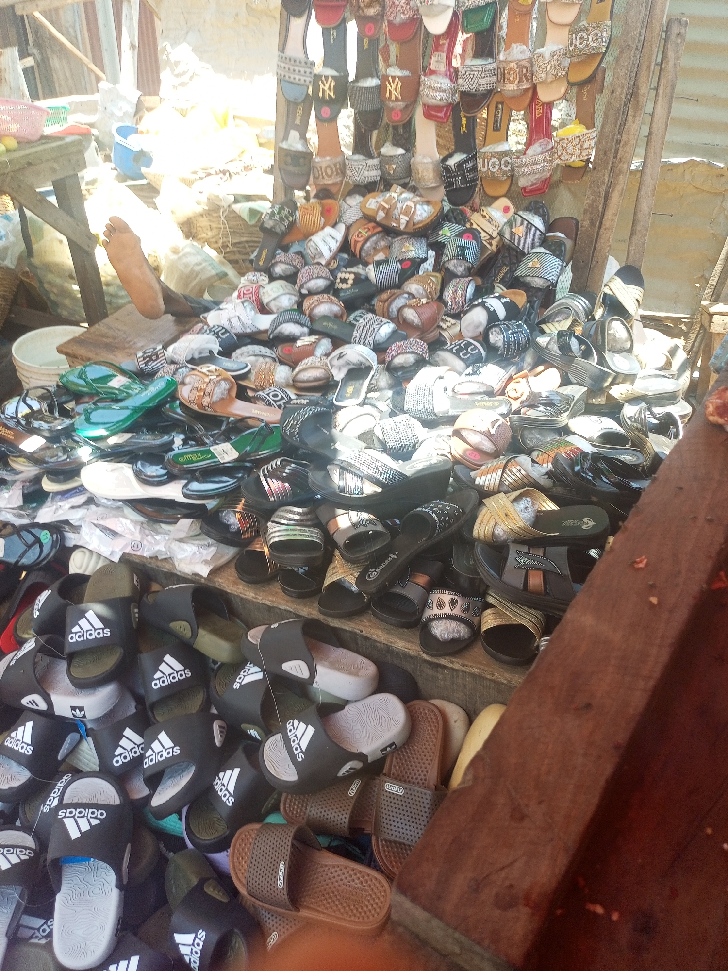 The image shows a vendor's stall in Sabon Tasha, Nigeria, piled high with a variety of sandals and slides. There are many pairs of slides with the "Adidas" logo, and many sandals of different styles, including some with designer brand names like "DIOR," "CELINE," and "GUCCI." The sandals are displayed on a tiered wooden stand. A person's bare foot is visible on a wooden structure in the background. The stall is shaded, likely for protection from the sun. The overall environment appears to be a bustling market setting.