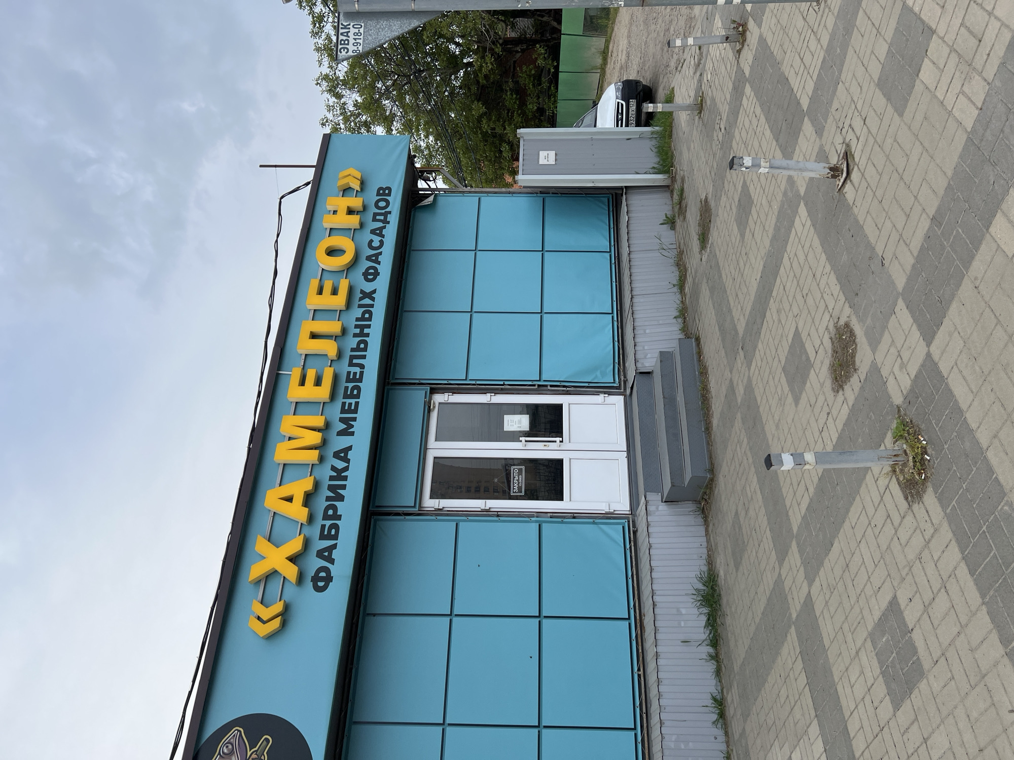 The image depicts a building facade in Krasnodar, Russia. The sign reads "Chameleon - Factory of Furniture Facades". The building is light blue with a rectangular grid pattern. The entrance is a white double door with a sign indicating it's closed. To the right of the entrance are steps. The sidewalk is made of gray and white pavers. There are three metal bollards. In the background, there is a tree, a gray structure, and a white car. A street sign is also visible in the background.