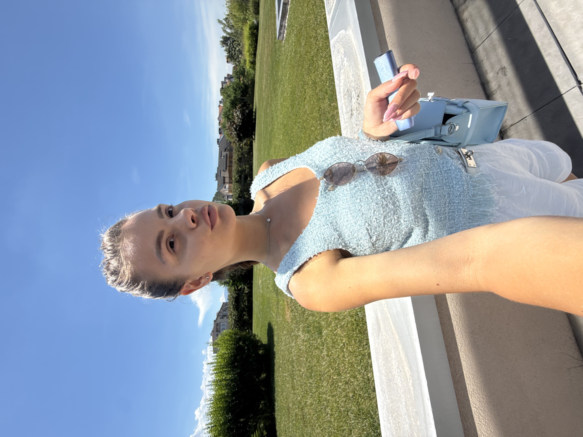 A young woman is captured in a selfie, her face tilted towards the camera. She is wearing a light blue, textured top and white shorts. A delicate silver necklace adorns her neck. Perched on the edge of her top are a pair of sunglasses. In her hand, she holds a light blue object, possibly a phone or a small device, and a matching light blue handbag rests beside her. Her fingernails are painted in a soft pink hue. The background reveals a lush green lawn bathed in the bright light of a sunny day, with trees and buildings visible in the distance, suggesting an outdoor setting. The clear blue sky dominates the upper portion of the image. The lighting indicates it is daytime, likely mid-morning or afternoon, with the sun casting shadows. The overall mood appears relaxed and casual, perhaps during leisure time.