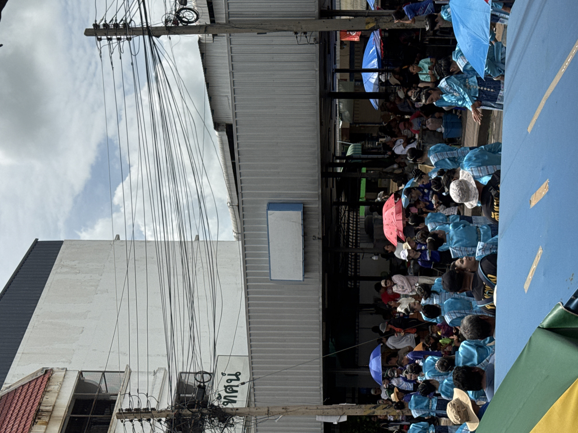 The image depicts an outdoor scene in Prakhon Chai District, Thailand, possibly during a festival or parade. A crowd of people, many wearing bright blue traditional outfits, are gathered. Some individuals hold umbrellas, one blue and two pink, providing shade. The background features a building with a red-tiled roof and white walls, along with a silver corrugated metal structure. A large cluster of electrical wires hangs prominently in the foreground, crisscrossing the scene. A sign with green lettering is visible on the white wall of the building. The overall impression is one of a lively community event.