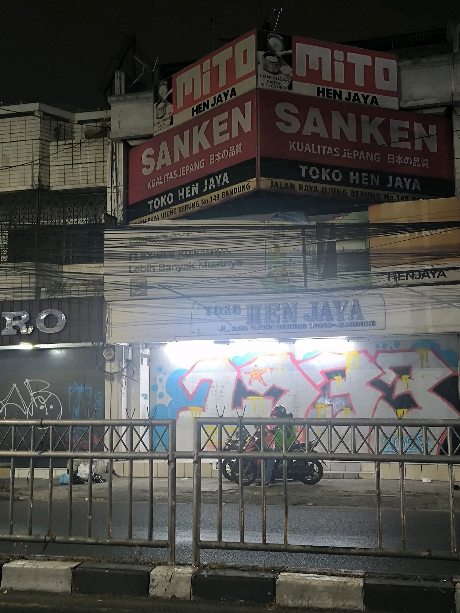This is a nighttime photograph taken in Bandung City, Indonesia, focusing on storefronts and street-level details. The scene is dominated by signage for electronics brands "Mito" and "Sanken," featuring slogans like "KUALITAS JEPANG" (Japanese Quality) and "TOKO HEN JAYA" (Hen Jaya Shop). Beneath these main signs, other banners and advertisements are visible, including one that reads "Lebih Banyak Muatnya" (More Capacity) and another with "RO".
The foreground is partially obscured by a metal fence with crisscrossing bars. Beyond the fence, a street is visible with a wet surface, suggesting recent rain or damp conditions. A motorcycle with a rider wearing a helmet is parked near a wall covered in vibrant graffiti featuring abstract shapes and lettering in pink, blue, and yellow. In the background, more buildings and electrical wires add to the urban, slightly chaotic atmosphere. The lighting suggests artificial light sources, consistent with a nighttime setting, casting shadows and highlighting the illuminated signs. The overall impression is of a commercial street at night, a mix of modern commerce and urban street art.
