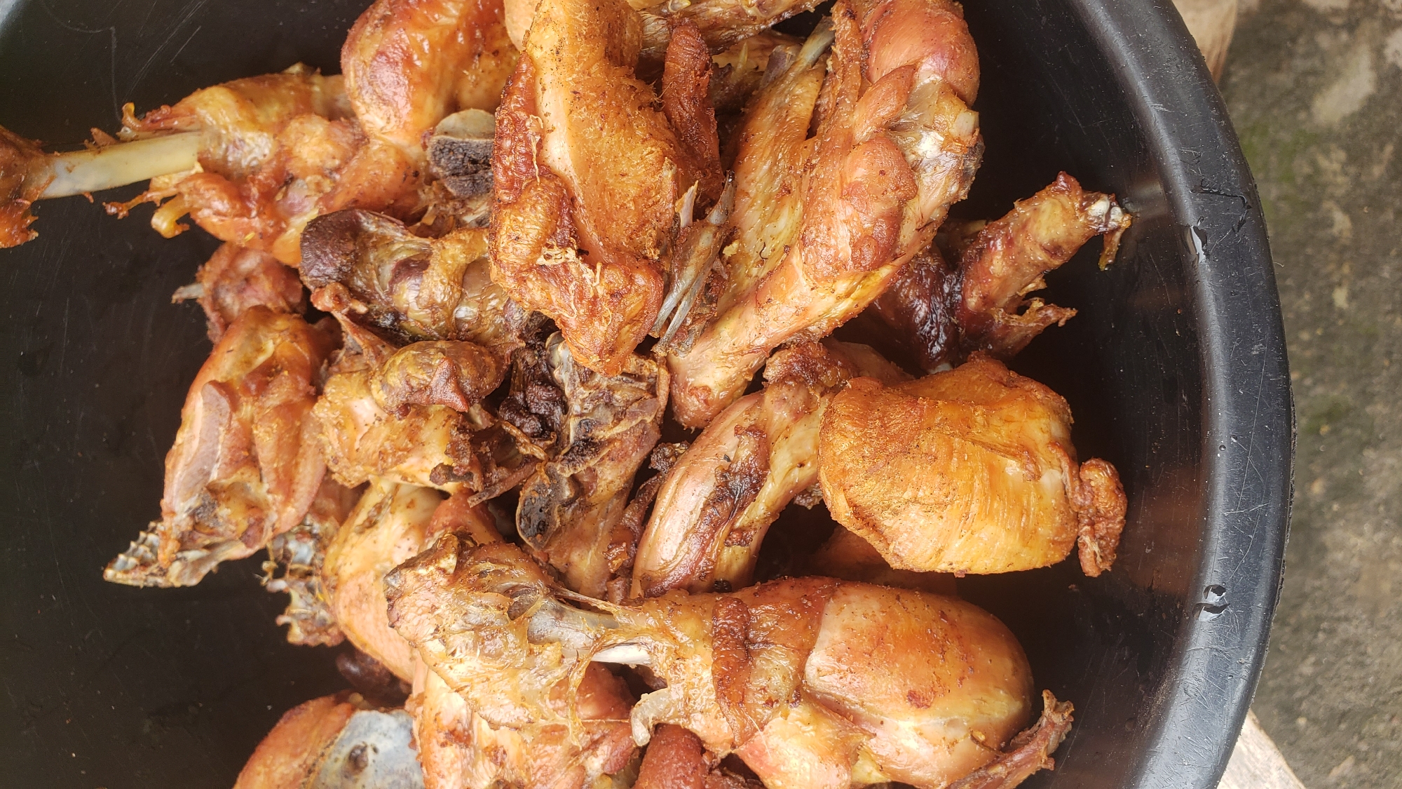 A close-up, overhead shot shows a large pile of golden-brown fried chicken pieces in a black plastic basin. The chicken pieces vary in size and shape, with some appearing to be drumsticks, wings, and other cuts. They are arranged densely, with some pieces overlapping others. The surface of the chicken appears crispy and slightly oily. The black basin's rim is visible at the bottom of the frame, with some water droplets on its surface. The background is dark and indistinct, suggesting an indoor or shaded outdoor setting. The scene is located in Sabon Tasha, Nigeria, where this preparation of fried chicken is a common dish.