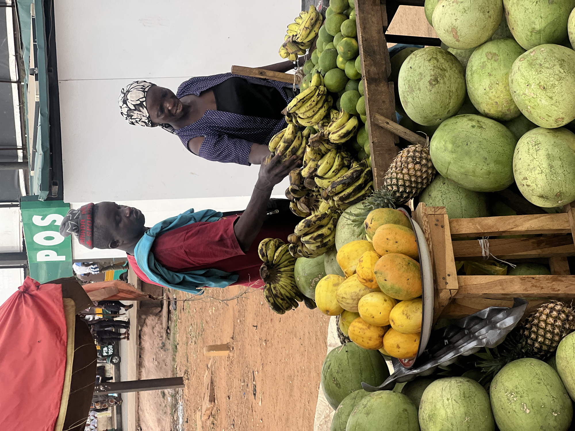 This vibrant image depicts an active outdoor fruit market stall in Gura Top, Nigeria, bustling with fresh produce and human interaction. Two individuals, a man and a woman, are the main subjects, seemingly engaged in commerce.
The woman, smiling gently, stands behind a wooden fruit display, her hands near a large cluster of ripe bananas. She wears a dark blue top with white polka dots and a black and white patterned headscarf. To her left, the man is visible from the chest up, clad in a red shirt with a blue garment draped over his shoulders and a patterned cap. He looks slightly towards the left side of the frame.
The market stall is abundantly stocked with a variety of fruits. Prominent displays include multiple bunches of yellow bananas, a large pile of green watermelons, and a wooden crate overflowing with green grapefruits or large oranges. A metallic bowl holds numerous yellow-skinned fruits, likely mangoes or papayas, and at least two pineapples are visible among the other fruits. The fruits are arranged on wooden crates and directly on the ground.
The background shows a dusty, reddish-brown ground characteristic of an outdoor setting. A red umbrella or awning provides shade, and blurred figures of other people and a vehicle with the text "PTA 0958" can be seen in the distance. A green sign on a building to the left prominently displays "POS," likely indicating a point-of-sale service. The scene is brightly lit, suggesting it is daytime.