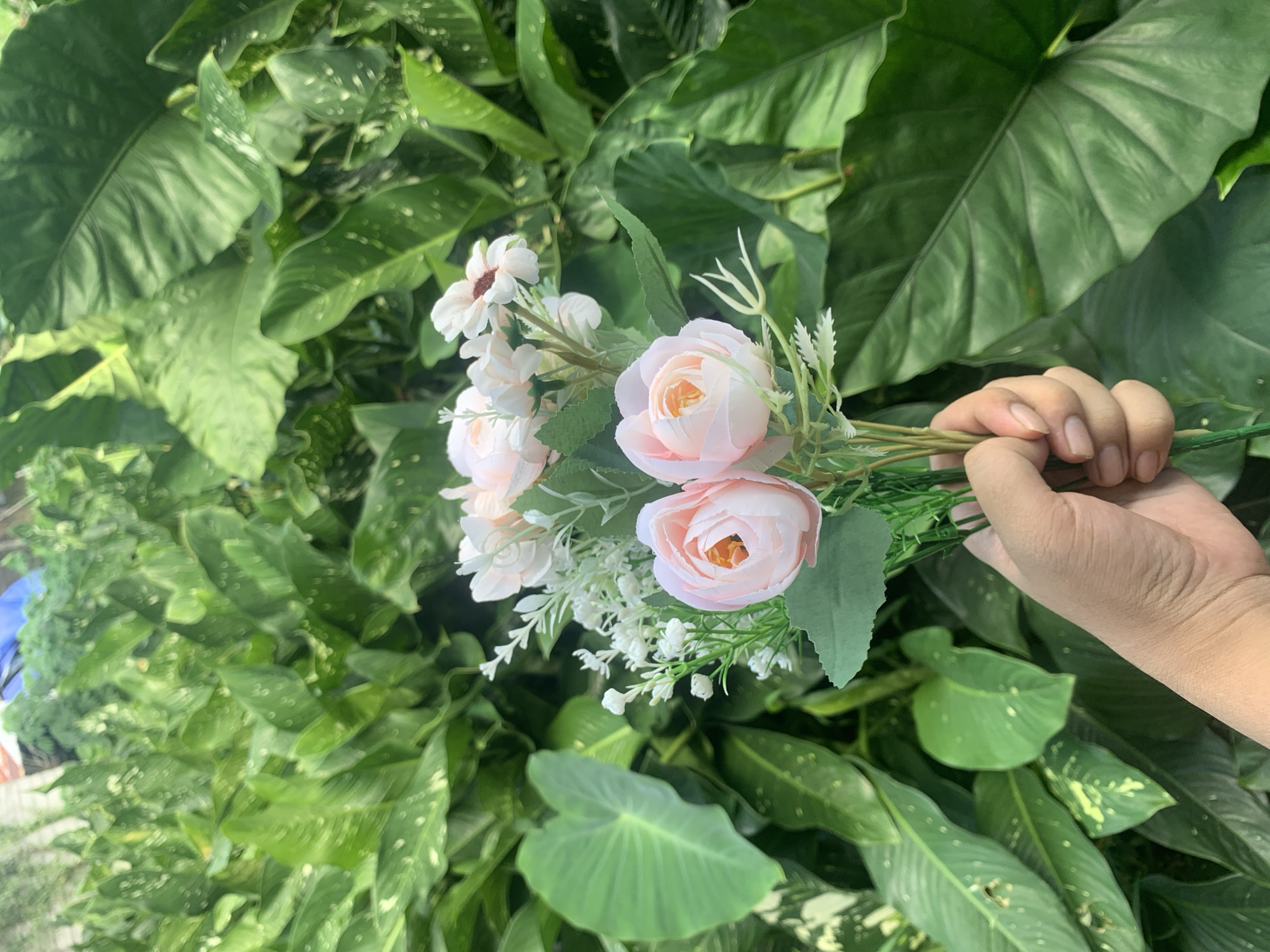 A person's hand, with light-colored skin and short fingernails, is holding a bouquet of artificial flowers. The bouquet consists of two pink roses with yellow centers, and several small white flowers with brown centers, along with green leaves and sprigs of greenery. The background is dominated by lush, dense foliage of various green plants, some of which have speckled patterns on their leaves. The overall scene suggests an outdoor setting, likely in a garden or natural environment, potentially within the city of Kuala Pembuang, Indonesia, known for its verdant landscapes.