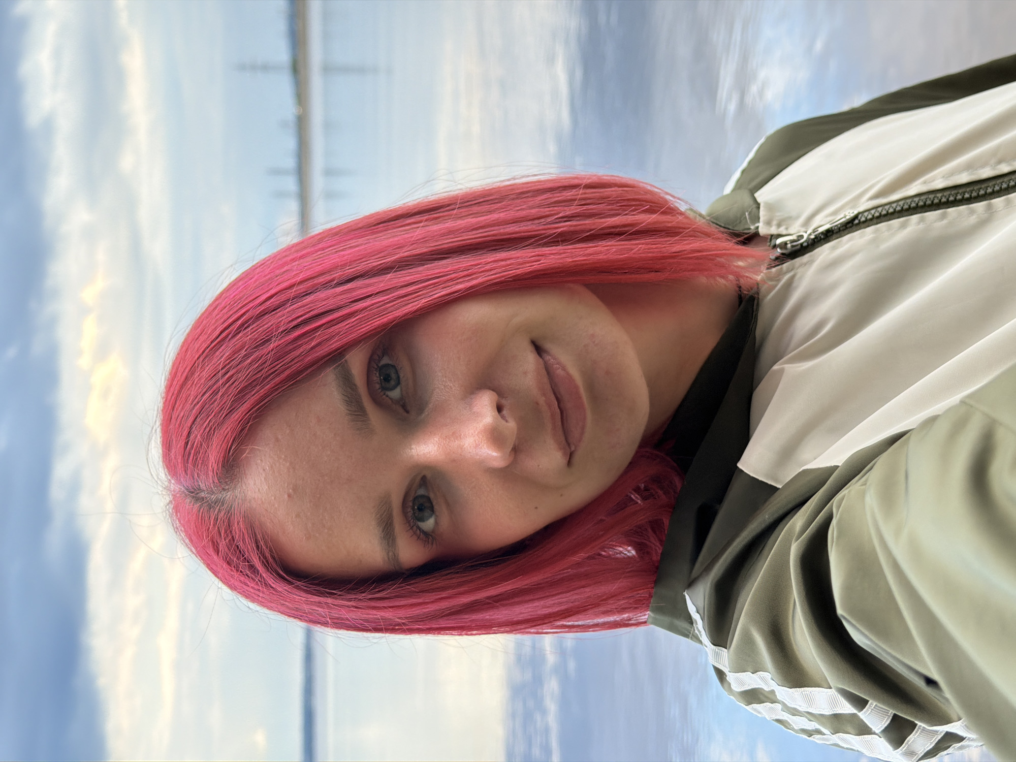 A young woman with vibrant pink hair takes a selfie outdoors, with a body of water and a cloudy sky in the background. She is wearing a green and white jacket with white stripes on the sleeves. The location appears to be Noviy, Russia. The water reflects the sky and clouds. In the distance, there are industrial structures visible.