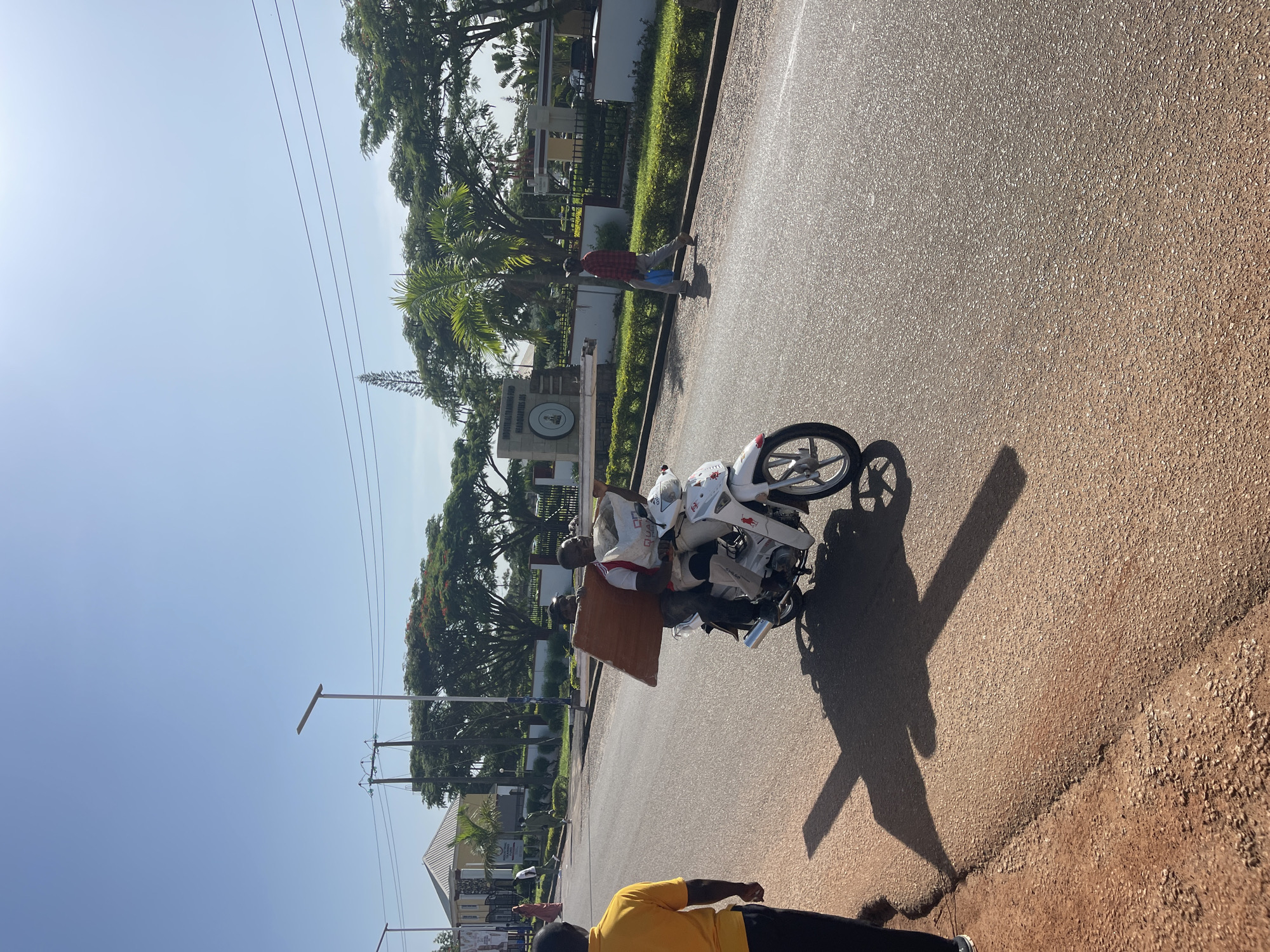 Two men are riding a white scooter on a paved road in Jos, Nigeria. The man in the back is holding a large wooden board. The man in the front appears to be wearing a red and white shirt and tan pants. The scooter has a white bag attached to the back. In the background, there are trees, a building with a white roof, and a sign that reads "INDUSTRIAL TRAINING FUND HEADQUARTERS." A man in a yellow shirt is walking on the side of the road, facing away from the scooter. The sun is shining, casting long shadows on the road.