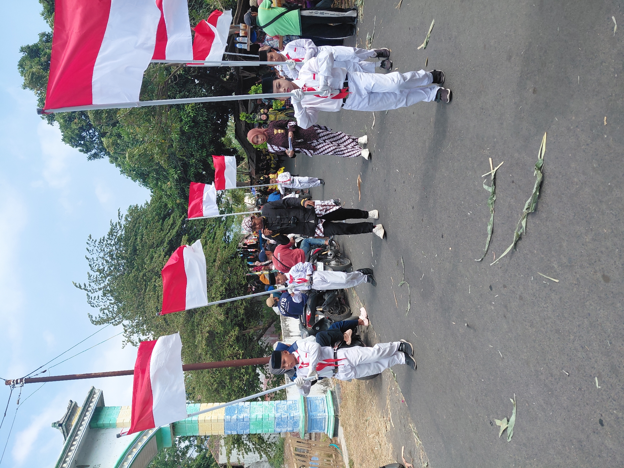 In Bimo, Indonesia, on a paved road under a bright sky, a procession is underway. Several children, dressed in white uniforms with red sashes, march in formation, each holding a flag pole. Some of the poles are topped with the red and white Indonesian flag, while others hold white flags. Behind them, adults also participate, some in traditional attire. One man in a black jacket and patterned pants plays a small instrument, likely a flute. Another woman wears a brown and white patterned dress and a headscarf. Motorcycles are visible in the background, with riders and passengers observing the procession. A decorative archway, painted in shades of blue, yellow, and green, stands on the left side of the road. The environment is somewhat rural, with trees lining the sides of the road and scattered debris, including leaves, on the asphalt.