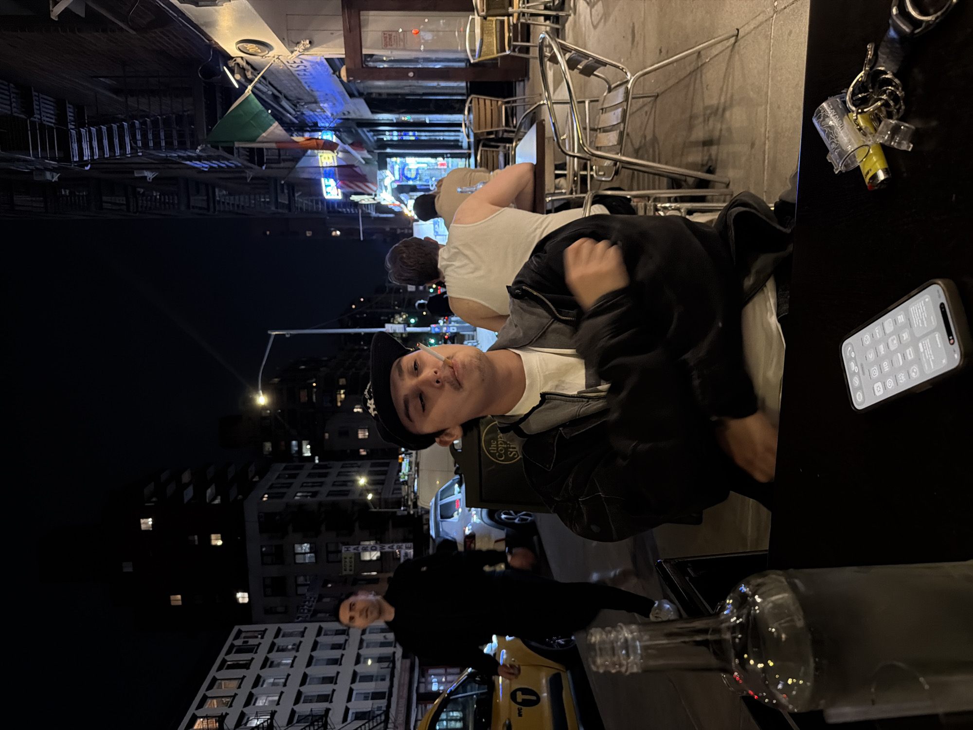 Here is a description of the image:
The image depicts an outdoor scene at night in New York City, United States. A person wearing a black baseball cap and a black jacket over a white shirt is seated at a table. A cigarette is visible in their mouth. Behind this individual, another person in a white tank top is also seated at the table.
Various objects are present on the table, including a smartphone, keys, and a partially visible glass bottle. The background features buildings with illuminated windows, streetlights, and an Italian flag hanging above the street, with other flags nearby. A taxi cab is visible in the lower portion of the image. Additional people are walking on the sidewalk in the background.