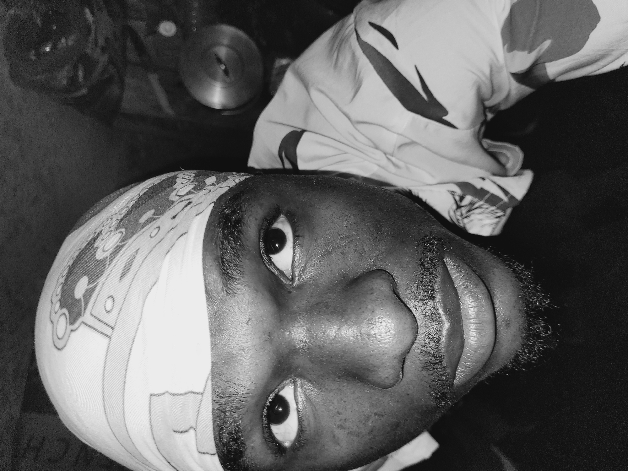 A close-up, black and white, high-angle shot shows a person wearing a bandana and a patterned shirt. The person is looking directly at the camera. The background is dark and out of focus, with some indistinct objects visible, including what appears to be a metallic circular object. The image is likely taken indoors, with the lighting creating strong shadows and highlighting the texture of the person's skin and facial hair. The bandana has a repeating pattern of stylized figures and geometric shapes. The shirt also has a bold, graphic pattern. No specific location details in Bukuru, Nigeria can be determined from the image itself.