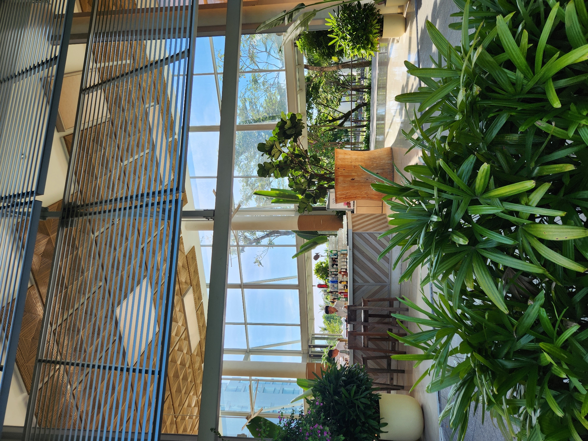 The image depicts the interior of a modern, open-plan hotel lobby or lounge area with large windows offering views of lush greenery and a bright blue sky, suggesting a tropical or subtropical climate. The scene appears to be set in Đà Nẵng, Vietnam, a coastal city known for its resorts and modern architecture.
In the foreground, dense, vibrant green foliage fills the bottom of the frame, creating a natural barrier that partially obscures the view of the interior. Behind this foliage, a contemporary bar area is visible, complete with a polished counter, bar stools, and an array of liquor bottles and glasses. Two individuals, appearing to be bar staff, are positioned behind the counter, one in a white shirt and the other in a light-colored uniform, engaged in what seems to be routine service. The bar is backed by a wall with a patterned design and large windows that open to an outdoor patio or garden area.
Further into the background, through the expansive glass windows, a well-maintained garden with various trees and plants is visible, along with glimpses of a building that could be a hotel or resort structure. The ceiling of the interior space is a prominent feature, with a modern design incorporating wooden elements and recessed lighting, contributing to the sophisticated ambiance. The overall impression is one of a tranquil, upscale environment designed to blend indoor comfort with outdoor beauty.