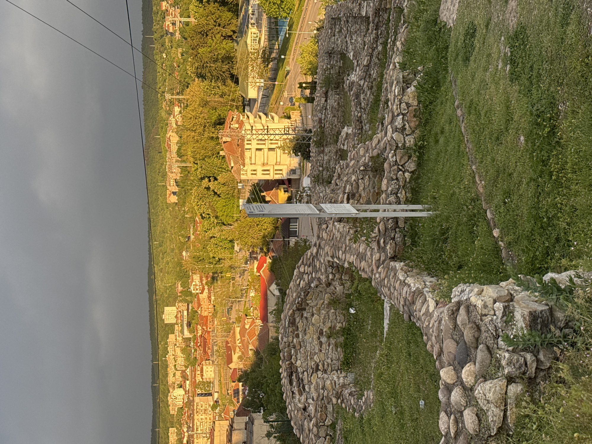 This image shows an elevated view of a city, likely taken from a historical site. The city is characterized by buildings with orange and red roofs, densely packed together and surrounded by trees. A road runs parallel to the city, with cars visible. In the foreground, there is a stone wall made of rounded rocks, presumably part of the historical site from which the photo was taken. The wall is covered in patches of grass. Two white signs are present, likely providing information about the site. In the upper left corner, there are power lines against a dark, cloudy sky, which contrasts sharply with the sunlit city below. The scene appears to be in the city of Unknown, Unknown.