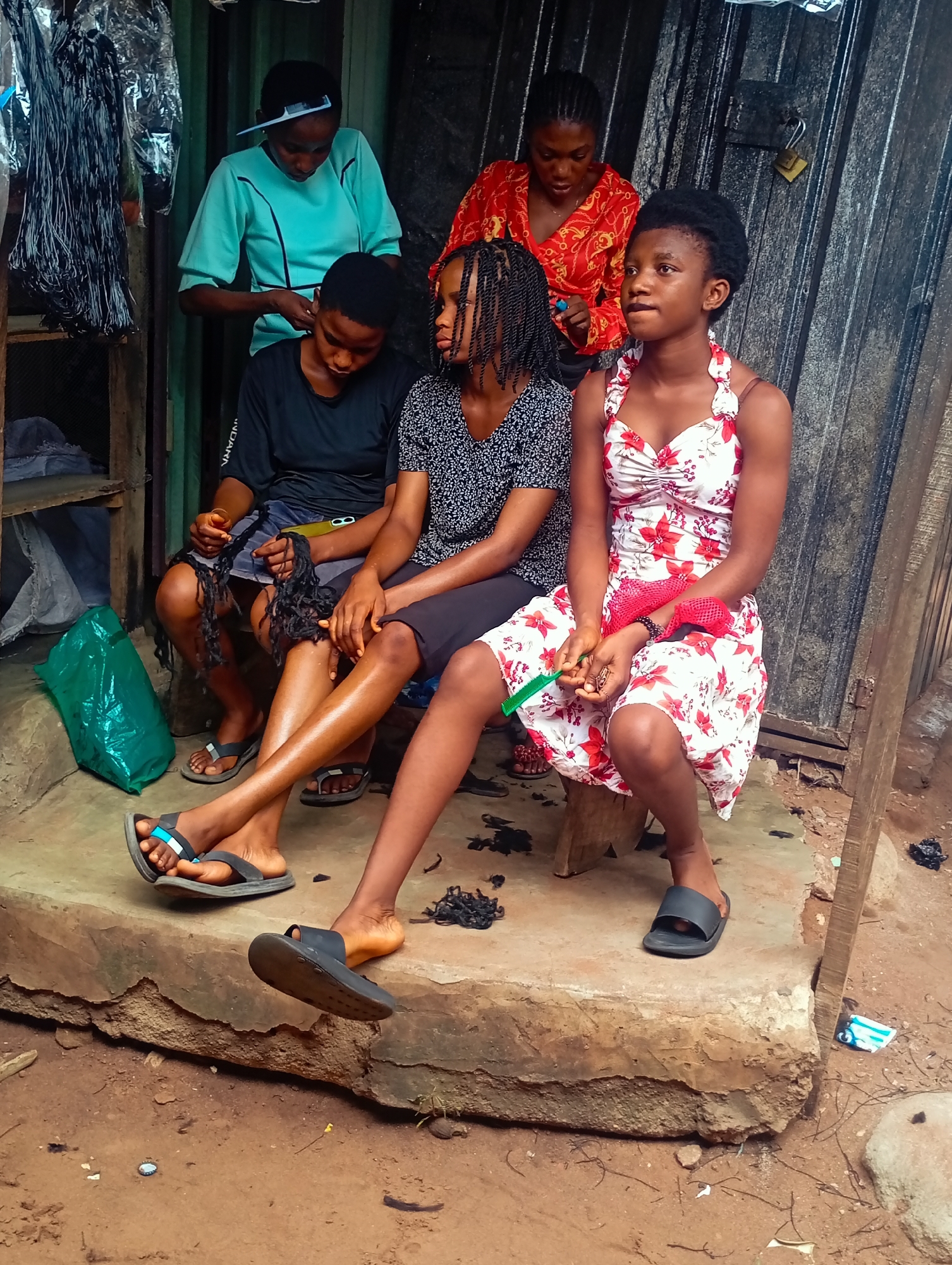 This media file depicts four young individuals, three females and one male, in what appears to be an outdoor setting, possibly a workshop or a roadside stall, in Nkpor, Nigeria. The individuals are engaged in hairstyling activities.
Two females are seated on a concrete step in the foreground. The one on the right wears a white dress with red floral patterns and a red accessory draped over her lap. She is holding a green comb. The female to her left, wearing a black and white patterned top, is also seated and appears to be having her hair braided by the person standing behind her. The third female, standing, wears a light blue top and is actively styling the hair of the seated female in the patterned top. In the background, another female, wearing a red and gold patterned top, is also present, looking towards the hairstyling action.
Objects visible include combs, hair strands scattered on the ground, a green plastic bag, and what appear to be bundles of hair or extensions hanging on the left side of the frame. The setting is rustic, with a corrugated metal structure in the background and a dirt ground. The lighting suggests it is daytime. The overall mood seems relaxed and communal, with the individuals focused on the task at hand. There is no visible text within the image.