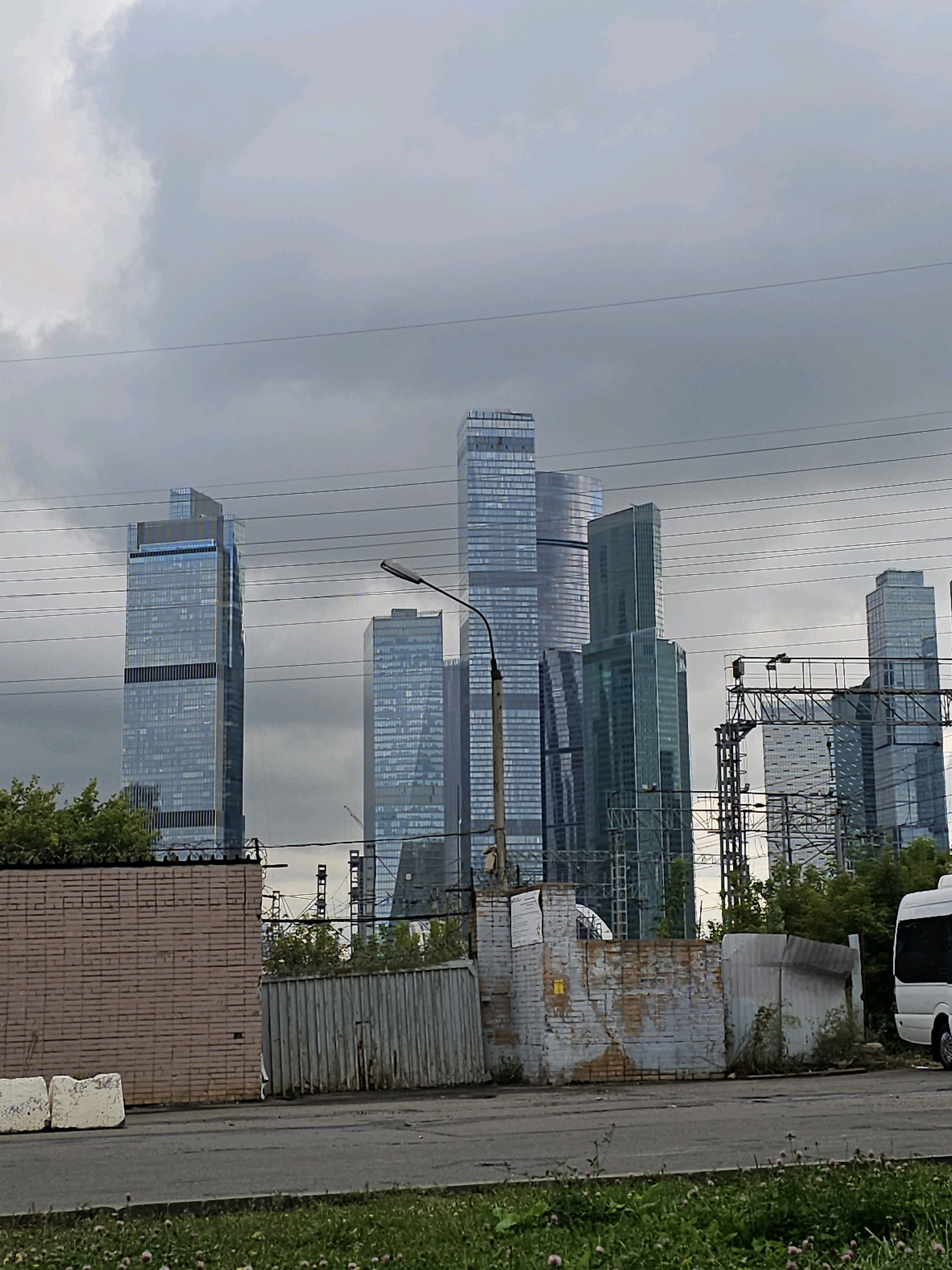 The image shows a view of the Moscow City business district under a cloudy sky. In the foreground, there is a paved road and a grassy area with some wildflowers. To the left, a weathered brick wall forms a barrier, with two large white concrete blocks in front of it. Next to the brick wall is a corrugated metal fence. In the background, several modern skyscrapers of varying heights and architectural styles dominate the skyline. One prominent skyscraper on the left is tall and slender with a blue-tinted glass facade. To its right are other glass skyscrapers, some with distinctive patterns. Overhead, power lines and a lamppost are visible. On the far right, the back of a white minibus is partially visible. The overall scene depicts an urban environment with a mix of industrial or utility structures in the foreground and a modern business district in the distance.