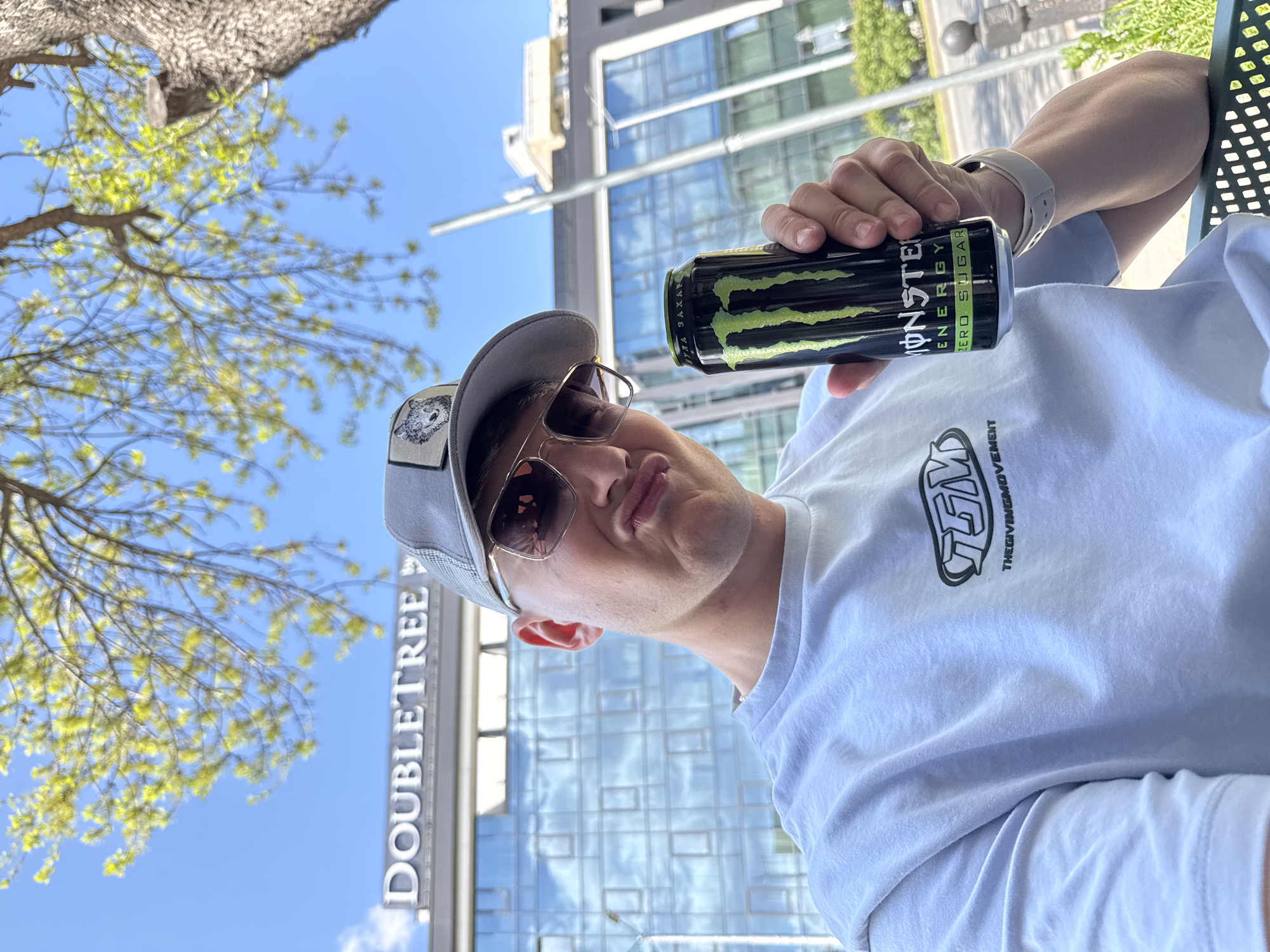 A man, presumably in his late teens or early twenties, is holding a can of Monster Energy Zero Sugar drink. He is wearing a grey baseball cap with a small patch featuring a bear design, sunglasses with reflective lenses, and a white t-shirt that has a logo of "TGM THEGIVINGMOVEMENT" printed. He's also wearing a light-colored smartwatch on his left wrist.
The background indicates that the man is outdoors. He is in front of a building with "DOUBLETREE" visibly displayed, suggesting it's a DoubleTree by Hilton hotel. The architecture behind him is modern, with a glass facade reflecting the sky. A tree with light green leaves is partially visible on the left side.
Based on the available information and visual context, the location is likely in Plovdiv, Bulgaria, specifically at or near the DoubleTree by Hilton Plovdiv hotel.