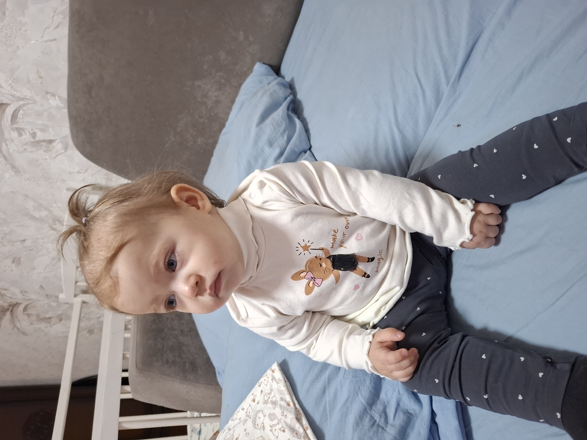 A baby is sitting on a blue bed in a room with a gray headboard and patterned wallpaper. The baby is wearing a white long-sleeved shirt with a rabbit design and gray pants with white hearts. The room appears to be in a residential setting, potentially in the city of Chaikovsky, Russia. The baby is looking off to the side with a serious expression. A white patterned blanket is near the bottom left corner of the image.
