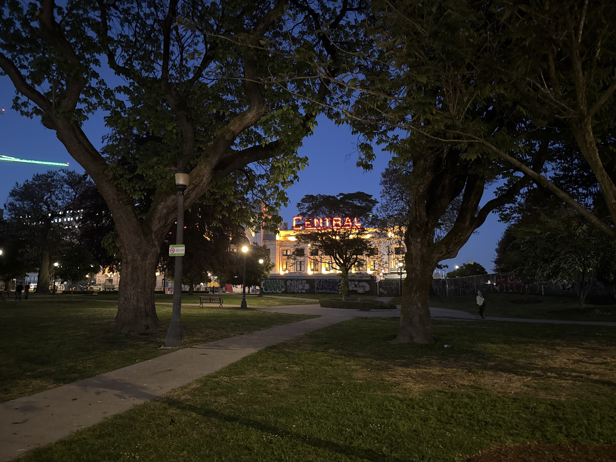 The image is taken at night in a park in Vancouver, Canada. Two large trees dominate the foreground, framing the view toward a building in the center, which is illuminated with an orange glow and features the word "CENTRAL" in large letters on its roof. The background displays the night sky, a gradient of blue hues. A paved path winds through the green grass of the park, leading toward the building. A lamppost with a small sign is visible to the left of the path. A fence with graffiti is visible behind the building. One person can be seen standing near the fence on the right. Benches are scattered throughout the park.