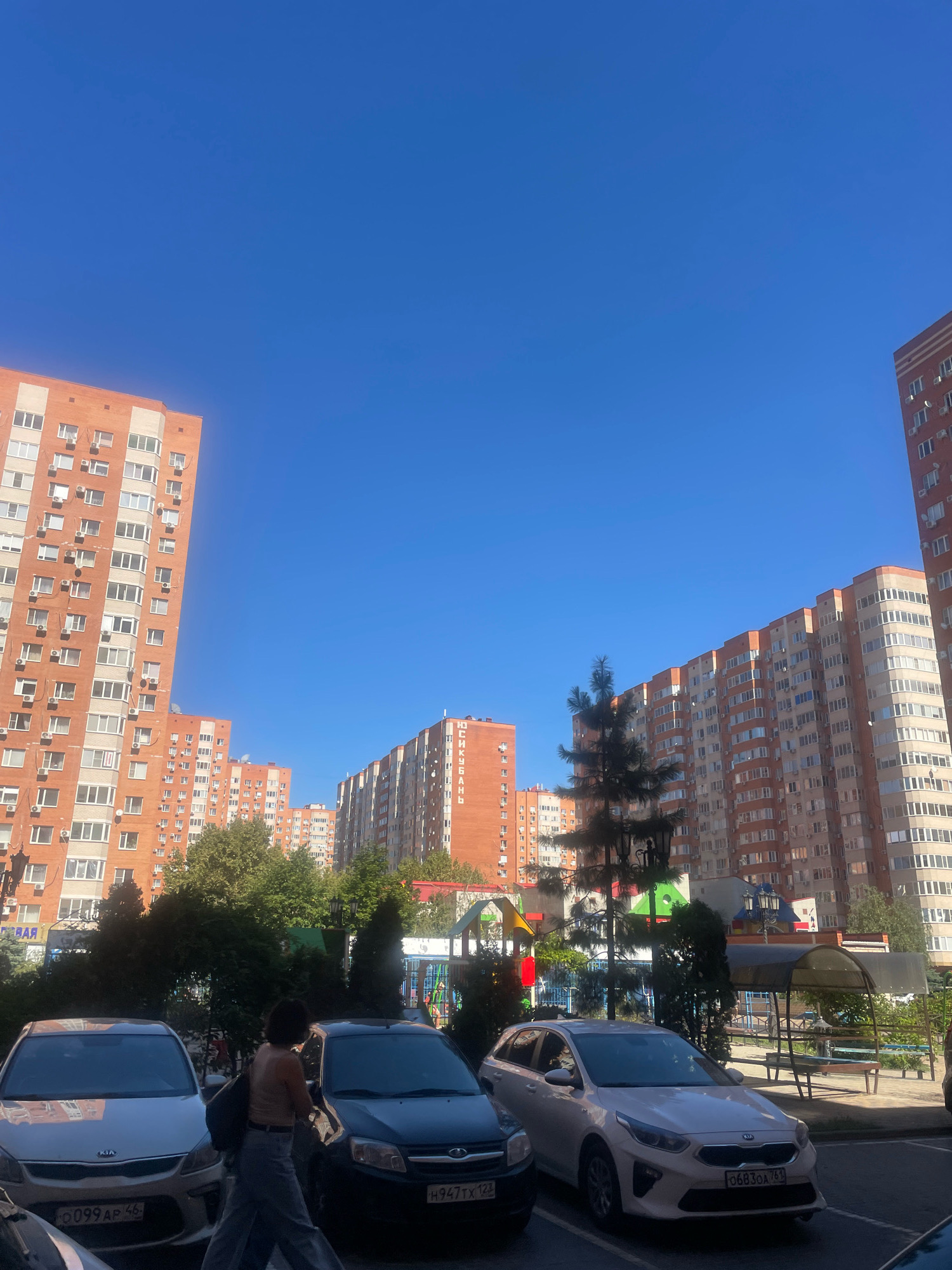 The image depicts a residential area in Krasnodar, Russia, on a clear, sunny day. In the foreground, several parked cars are visible, including a silver Kia Rio and a dark sedan. A woman with dark hair, wearing a light-colored sleeveless top and grey pants, walks between two of the cars, carrying a dark bag over her shoulder. Behind the cars, lush green trees partially obscure a playground area with colorful structures. In the background, tall, modern apartment buildings dominate the skyline. These buildings are orange and white, with numerous windows and air conditioning units visible. The sky is a vibrant, cloudless blue. One of the buildings has Cyrillic lettering that appears to spell "ЮЖКУБАНЬ" (Yuzhkuban). The scene suggests a typical urban residential environment with amenities like a playground and ample parking.