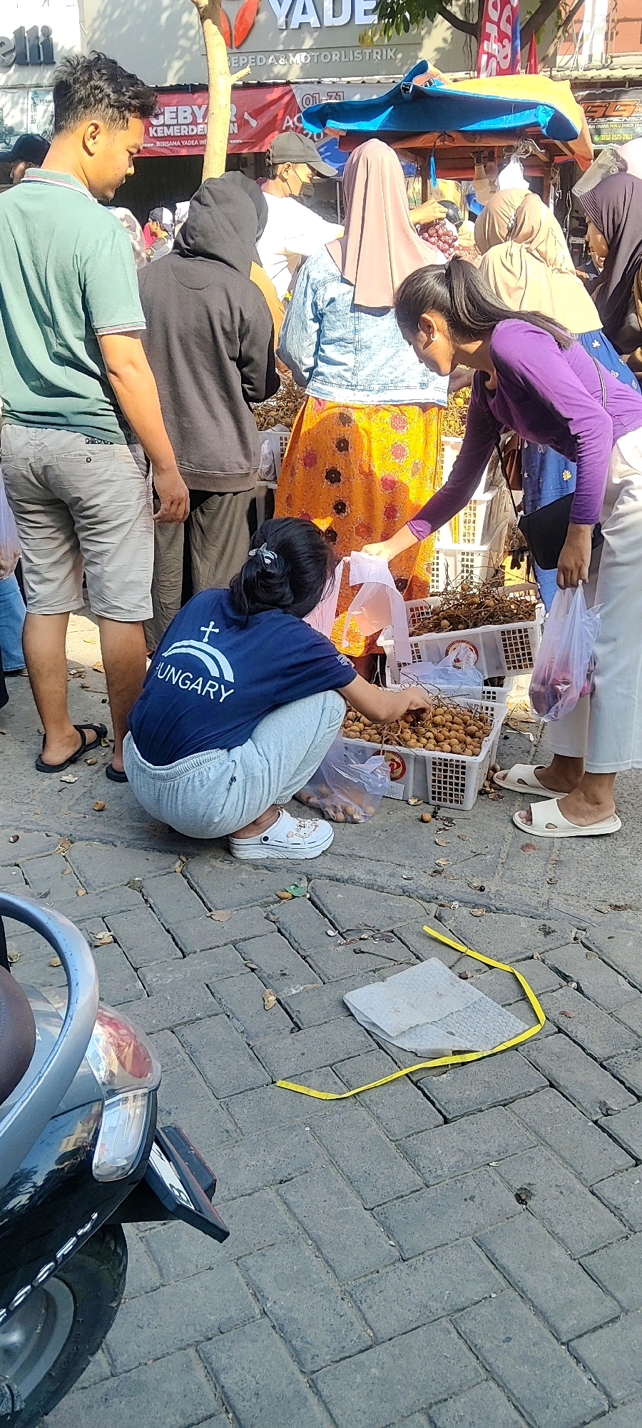 A medium shot captures an outdoor scene in Cibatu, Indonesia, likely a market or street vendor area, with a focus on people interacting around a fruit stall. In the foreground, a person wearing a dark blue shirt with "HUNGARY" written on it is crouched down, reaching into a white plastic crate filled with small, round, brown fruits. They are wearing light gray sweatpants and white clogs. To the right, a person in a purple long-sleeved shirt is extending a transparent plastic bag, possibly to receive fruit or to hand something to the crouched individual. This person is wearing white trousers and white slip-on sandals. Behind them, another person in a denim jacket and a patterned orange skirt stands, and further back, a person in a beige hijab and another in a dark hooded jacket are visible. To the left, a person in a green t-shirt and light gray shorts stands, facing away from the camera. The ground is paved with grey rectangular bricks, with some fallen leaves and debris scattered around. A dark-colored motorcycle is partially visible in the bottom left corner of the frame. In the background, a market stall structure with a blue and yellow canopy is visible, along with signage that appears to be in Indonesian. The overall lighting suggests it is daytime with bright sunlight.