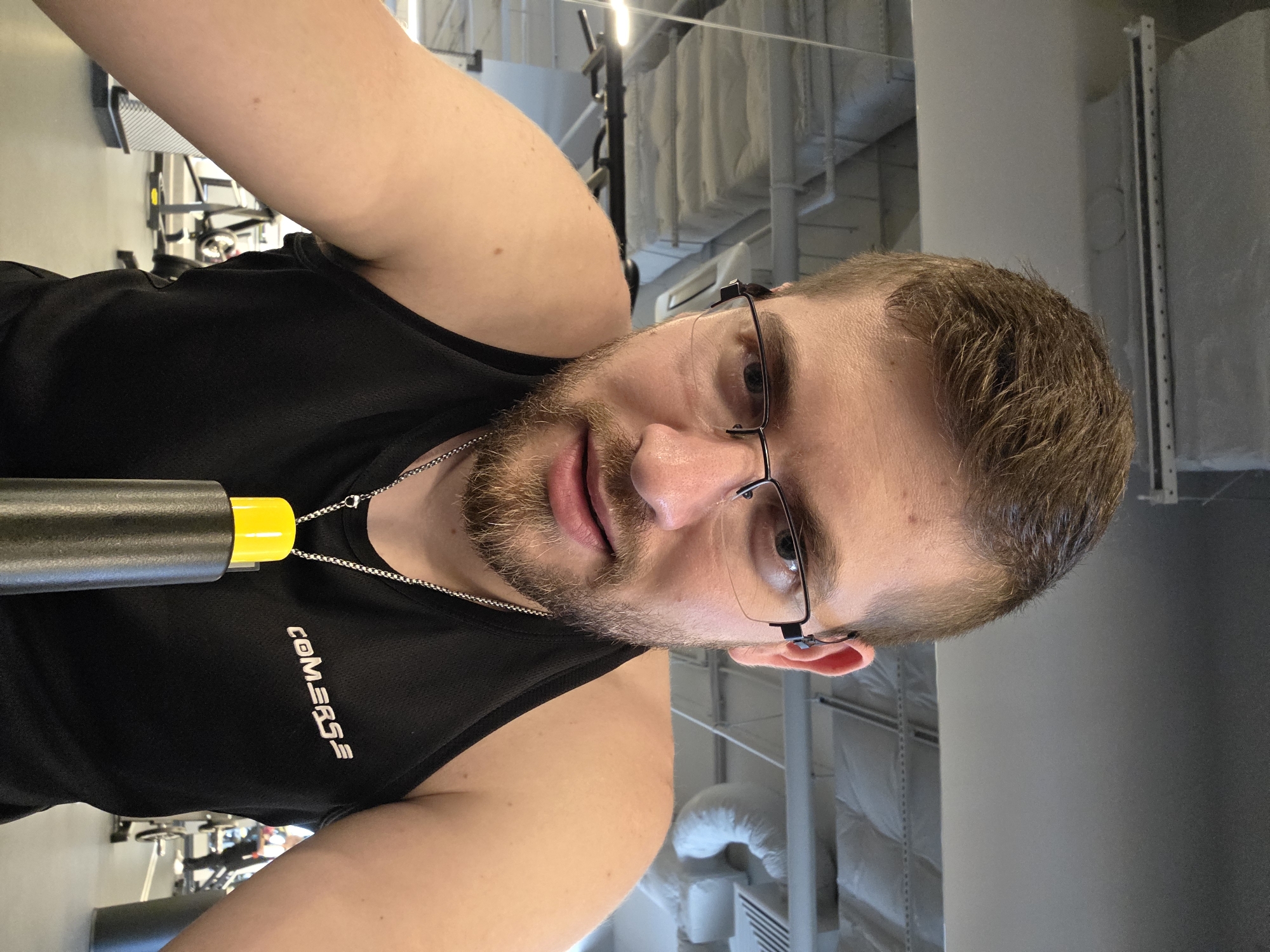 This image captures a close-up, slightly rotated selfie of a man inside a gym, likely located in Plovdiv, Bulgaria. The man, appearing to be in his late 20s or early 30s, has short brown hair and a neatly trimmed beard. He is wearing clear, black-framed glasses and looks directly at the camera with a neutral expression.
He is dressed in a black sleeveless athletic shirt, prominently displaying the white text "CONVERSE" vertically on the left side of his chest. A silver chain is visible around his neck. To the left of his torso, a portion of a black object with a bright yellow cap is visible, possibly a water bottle or part of a piece of gym equipment.
The background is out of focus but clearly depicts a gym setting with various exercise machines and equipment. Overhead, industrial-style ceilings with exposed white pipes and ventilation ducts can be seen, characteristic of a modern fitness facility. The lighting is artificial, suggesting an indoor environment. The image seems to capture a moment during or after a workout session.