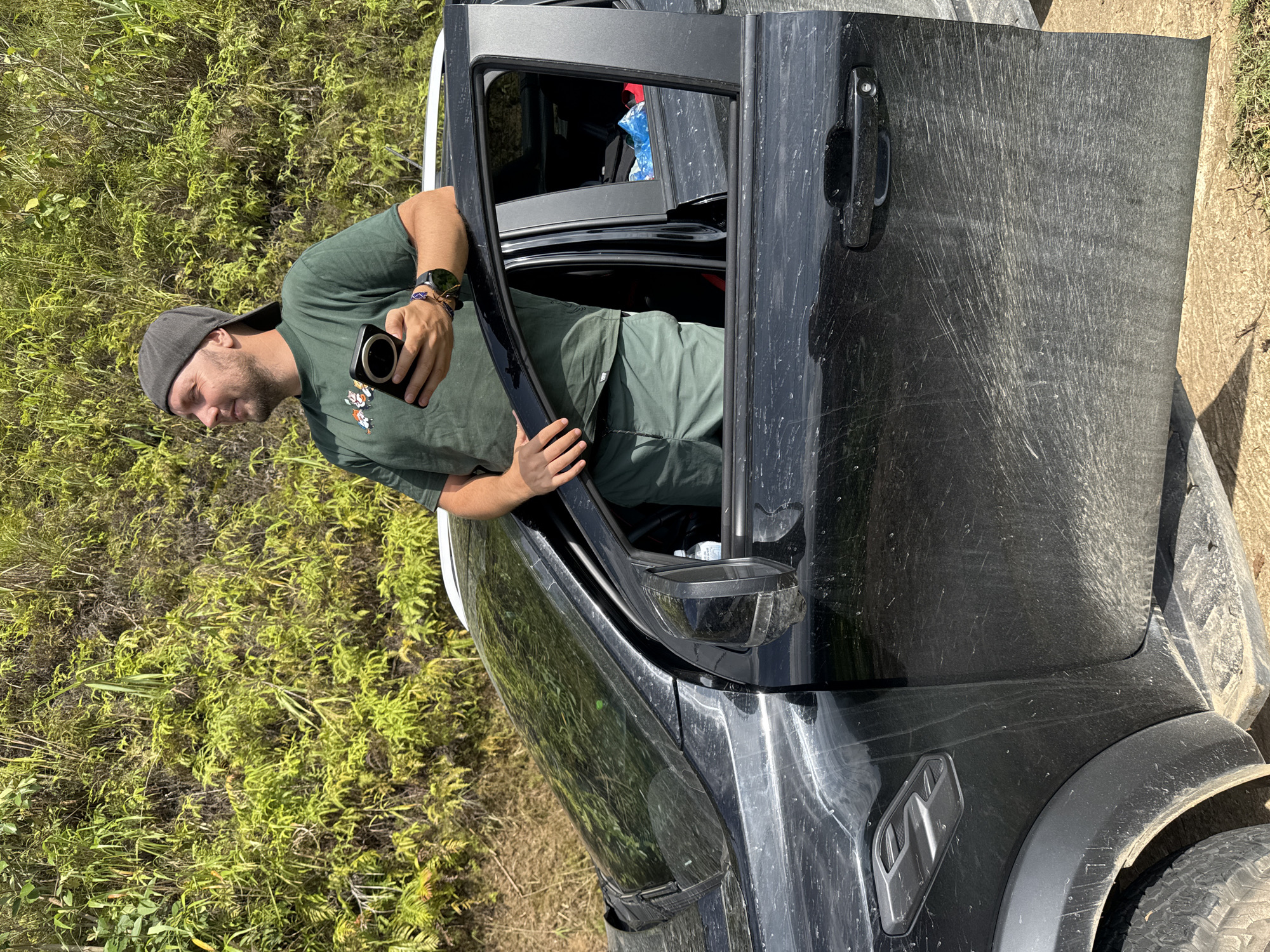 An outdoor, brightly lit image, taken in what appears to be a rural or off-road setting in Khao Mang, Vietnam, shows a man leaning out of the driver's side door of a heavily dirt-splattered and scratched black truck or SUV. The time of day is mid-day, indicated by strong shadows and clear sunlight.
The man, with a short beard and wearing a dark green t-shirt with a graphic of ducks/geese, a dark cap, and dark pants, is holding a black smartphone with a prominent multi-lens camera module. He has a watch on his left wrist and beaded bracelets. His gaze is directed downwards, suggesting he is using the phone's camera, possibly taking a photo or video of something below or beside the vehicle. His right hand rests on the truck's door frame.
The vehicle itself is robust, evidenced by its dark color, large tires, and significant accumulation of dirt and scratches on its bodywork, particularly noticeable on the open door. This condition strongly suggests recent off-road travel. The interior of the truck is partially visible through the open door, showing some red fabric inside.
The background consists of dense, lush green vegetation, including ferns and various grasses, indicating a verdant, possibly tropical environment. The ground around the vehicle appears to be a dirt track. There is no discernible text visible in the image.