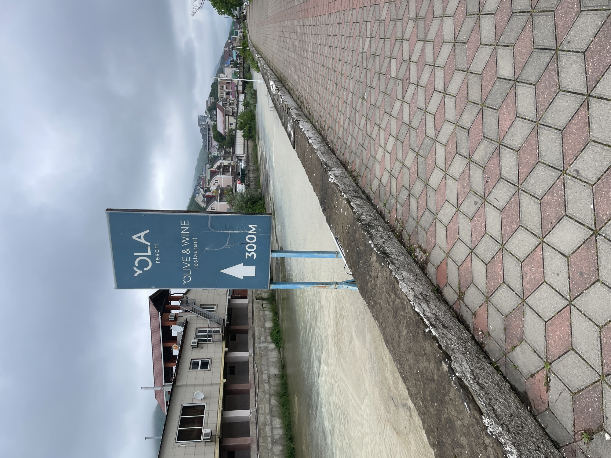 A sideways, eye-level shot shows a sign in Olginka, Russia. The sign reads "YOLA resort OLIVE & WINE restaurant" with an arrow pointing forward and "300M" underneath. It's located on a small concrete structure, likely a walkway, next to a body of water. The walkway is paved with a patterned brick design in gray and rose tones. Across the water is a building with multiple windows, a brown roof, and several air conditioning units. In the background, there are houses built on a slope. The sky is overcast and gray.