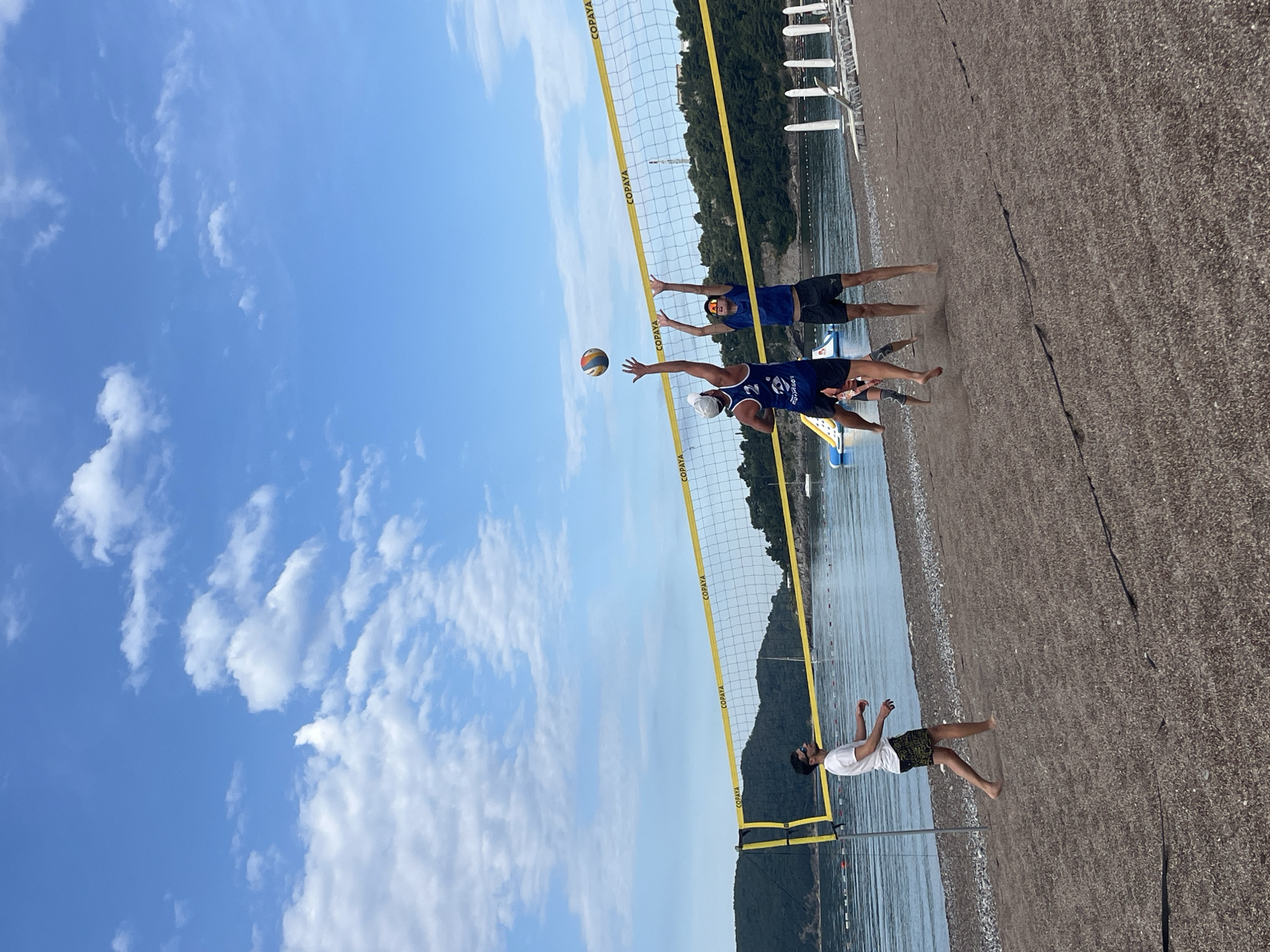 The image captures a beach volleyball game in progress. Two players, wearing blue shirts and dark shorts, are positioned near the net, with one player in the foreground actively hitting the volleyball. A third individual, wearing a white shirt and patterned shorts, is situated further back on the sand. The volleyball net, marked with the word "COPAVA," divides the players, and the backdrop consists of a beach, water, and greenery-covered land. The sky is blue with scattered clouds. The scene is likely set in the city of Boreti, Montenegro.