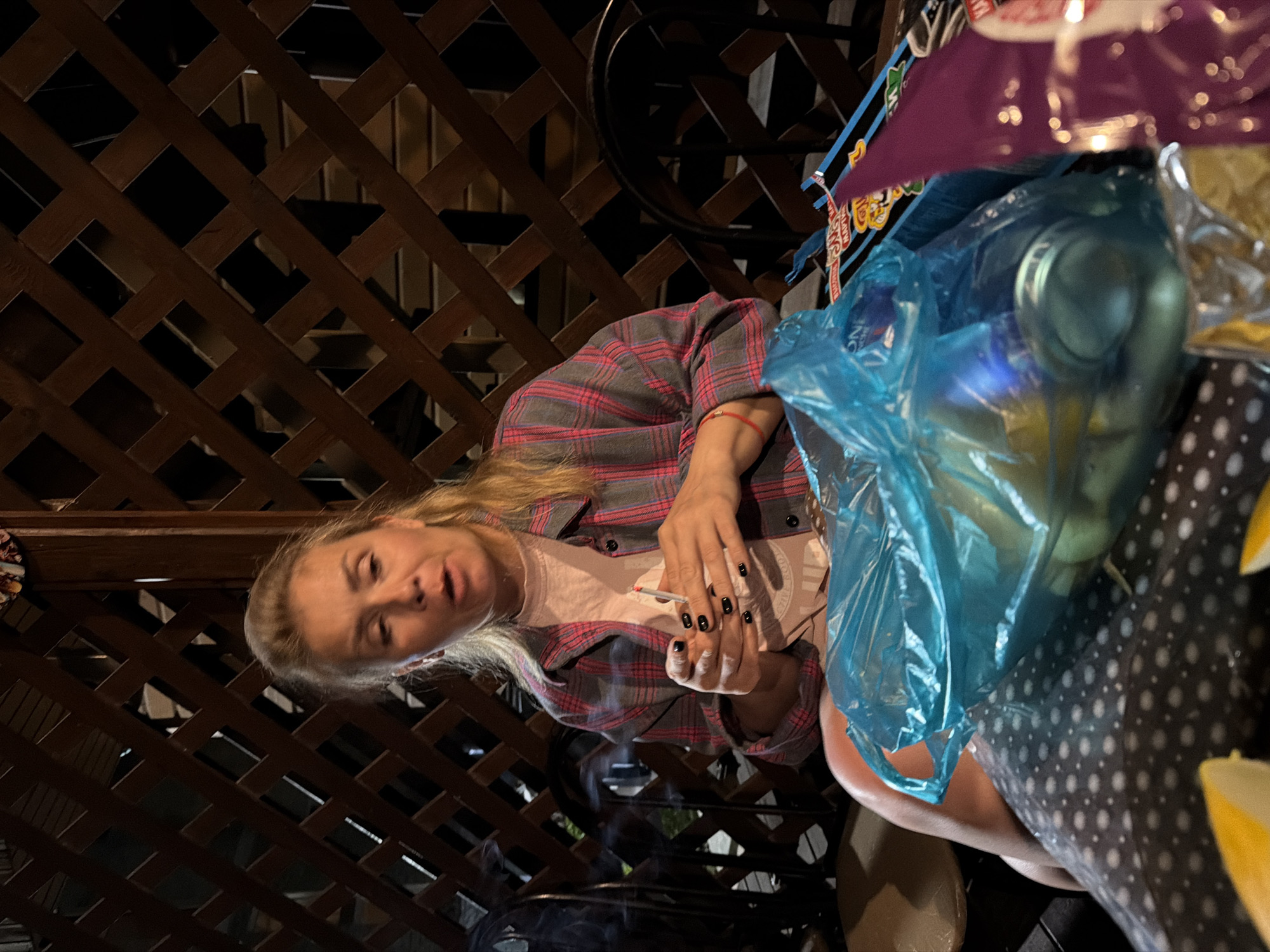 An outdoor scene at night in Sochi, Russia, features a woman sitting at a table on a patio. She has blonde hair, is wearing a plaid shirt over a pink t-shirt, and is holding a cigarette with a lit end and smoke rising from it. Her fingernails are painted black. In front of her is a blue plastic bag containing a light-colored object, possibly food, and on the table are wrappers of snacks. The background consists of a wooden lattice fence.