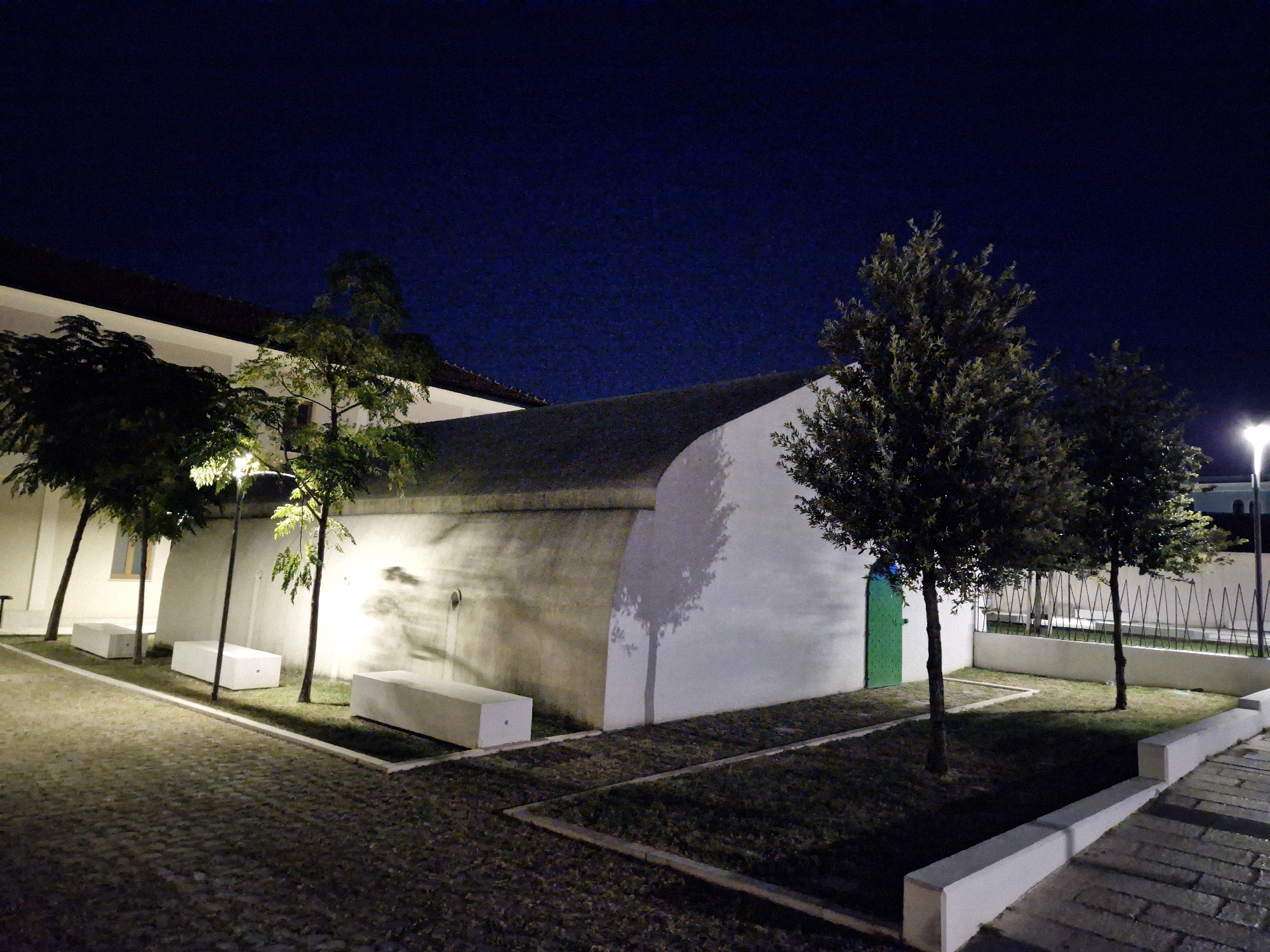 The image depicts an outdoor scene at night, illuminated by artificial lighting. In the foreground, a cobblestone pathway leads to a landscaped area with grass and trees. Several white, rectangular benches are placed in the grassy sections.
Dominating the mid-ground is a long, curved, white building with a dark roof. Its wall is illuminated, casting shadows of trees onto its surface. A green door is visible on this building. Adjacent to this building is another, lighter-colored building with a dark roof, partially visible on the left side of the image.
In the background, more trees are present, and a tall, white lamppost is illuminated on the right. The sky is a deep, dark blue, suggesting it is late evening or night.
The scene is located in the city of Taranto, Italy. Specifically, the area resembles the courtyard or garden of the Museum of Taranto (MARTE), located within the former St. Pasquale Baylon church complex in the historic center. The architectural style of the curved building and the presence of benches and landscaping are consistent with the museum's outdoor spaces designed for visitor engagement and events.