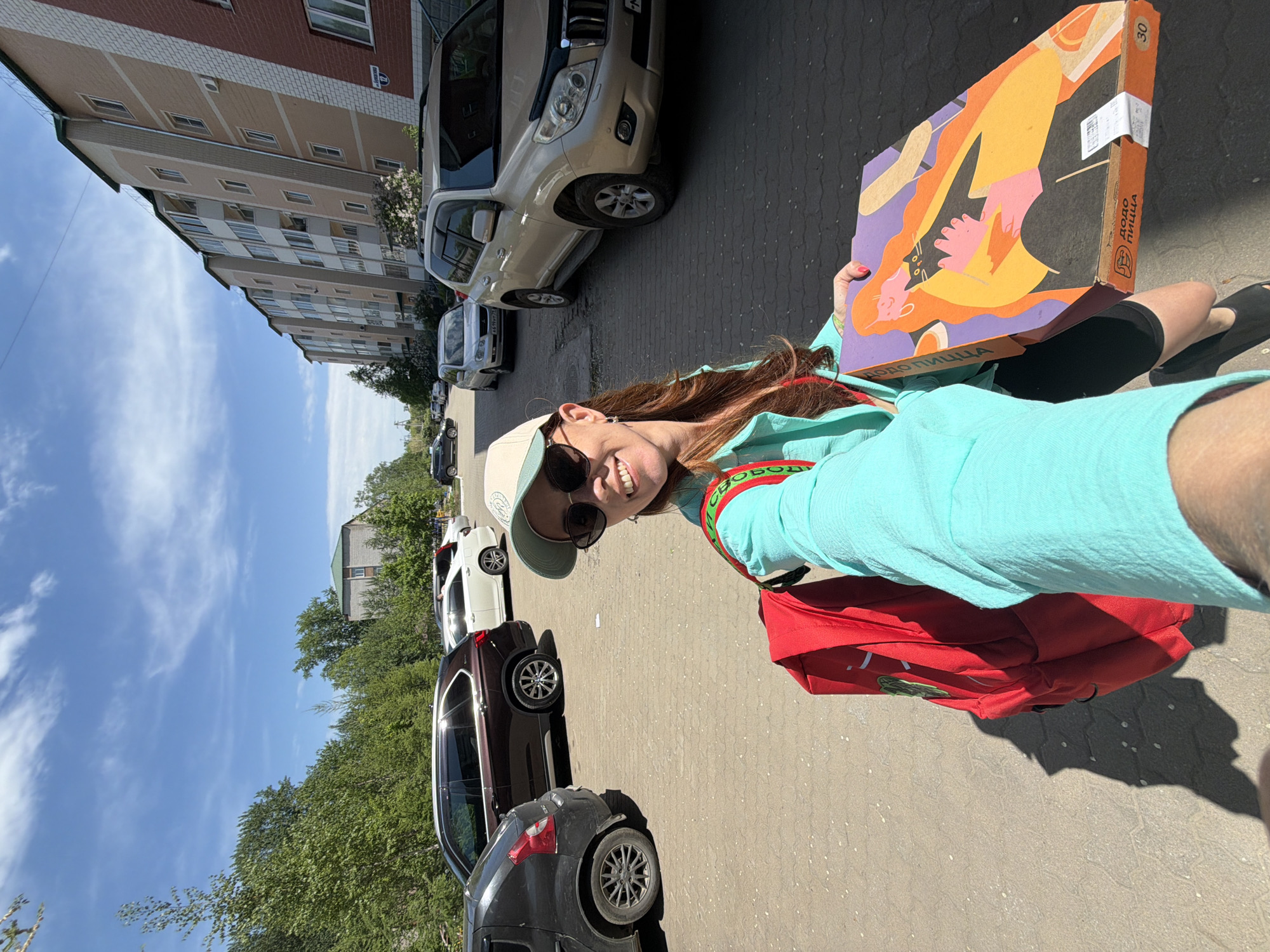 A woman stands in a parking area, holding a pizza box with graphic art, likely in Birobidzhan, Russia. She wears sunglasses, a cap, and a light blue shirt. A red bag hangs from her shoulder. The parking area includes several cars parked in a row. Buildings and trees are visible in the background, beneath a blue sky with sparse clouds. "Dodo Pizza" is printed on the box. The box also displays the number "30."