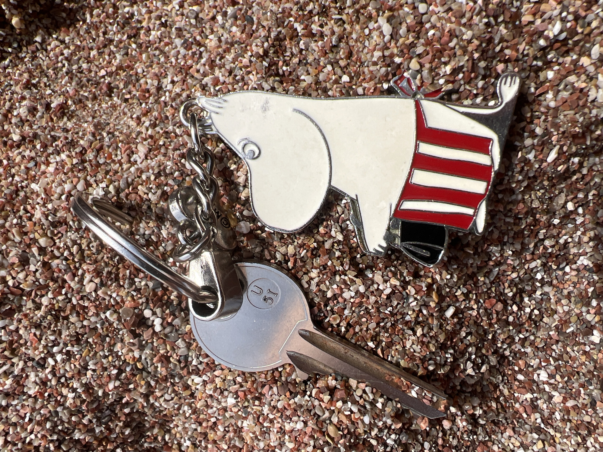 A keychain and a single key rest on a surface of small, reddish-brown and light-colored pebbles, characteristic of a beach or rocky shore. The keychain features a white, Moomin-like character with a distinctive snout and round ears, wearing a red and white horizontally striped outfit. The character's form is outlined in silver metal, with matching silver chain links connecting it to a keyring. Attached to this keyring is a single, silver-toned metal key. The key's head is circular, with the alphanumeric inscription "U 1C" clearly embossed on its surface. Bright, direct sunlight illuminates the scene, creating strong highlights on the metallic elements and distinct shadows on the pebbles. The setting is outdoors, suggesting a sunny day in a coastal area, specifically Petrovac centar, Montenegro, as per the provided context. There is no specific activity depicted, only the static arrangement of the objects on the ground.