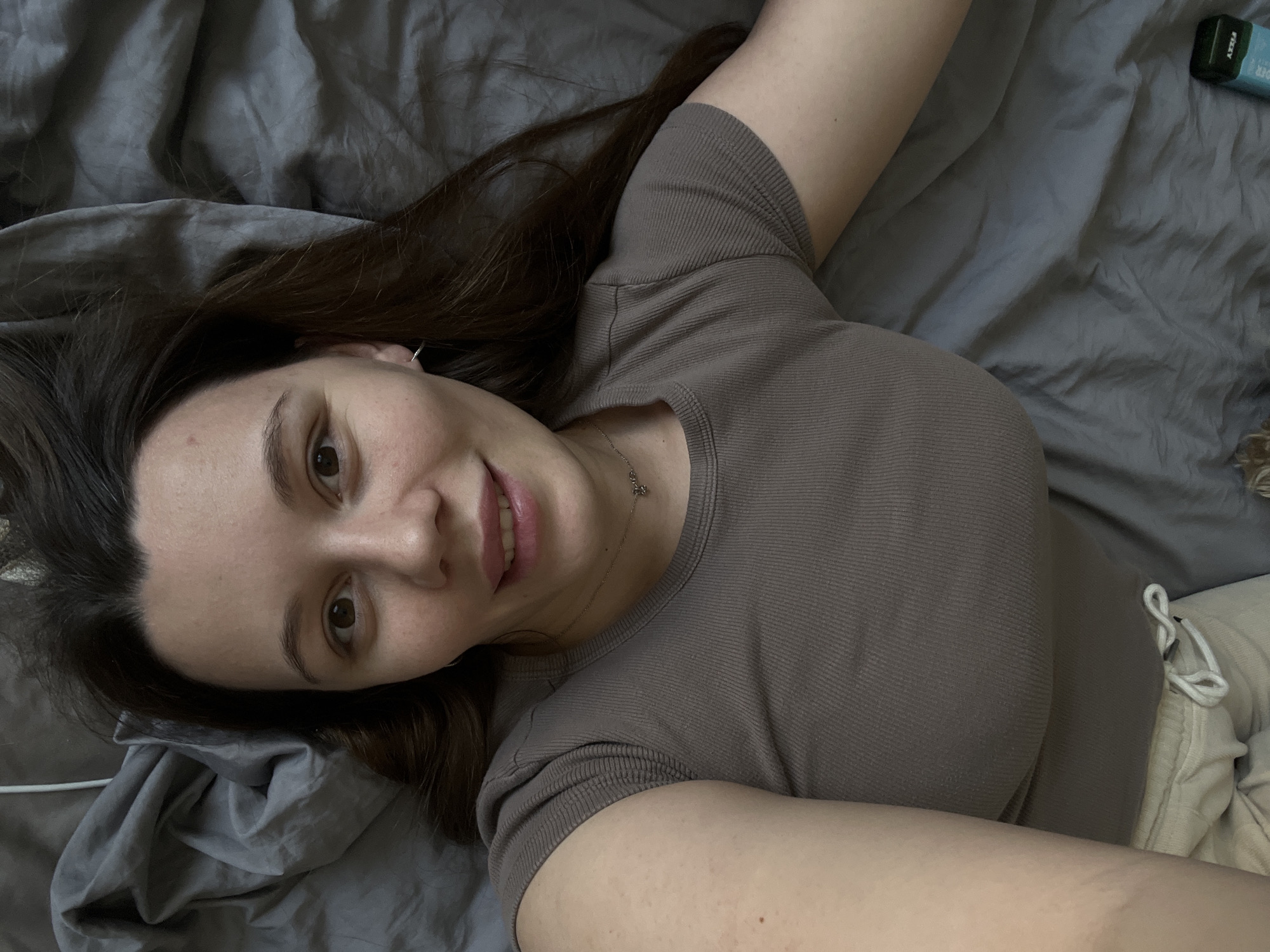 A single young adult female is depicted lying supine on rumpled gray bedding, viewed from a top-down perspective, indicative of a self-portrait. She has dark brown hair, brown eyes, and a light complexion, exhibiting a direct gaze and a slight smile. She wears a short-sleeved, ribbed, taupe-colored shirt, with light-colored, drawstring-style lower garments partially visible. A delicate silver-colored chain necklace with a small pendant is around her neck, and a small silver hoop earring is present in her visible left ear. A white cable is visible on the bedding in the upper left. In the lower right, a small, rectangular object with a light blue label (partially legible "FIZZY" and "AXXYA") and a dark green top rests on the bedding. The scene is indoors, offering no visual indicators for the specified city of Leninakan, Russia.