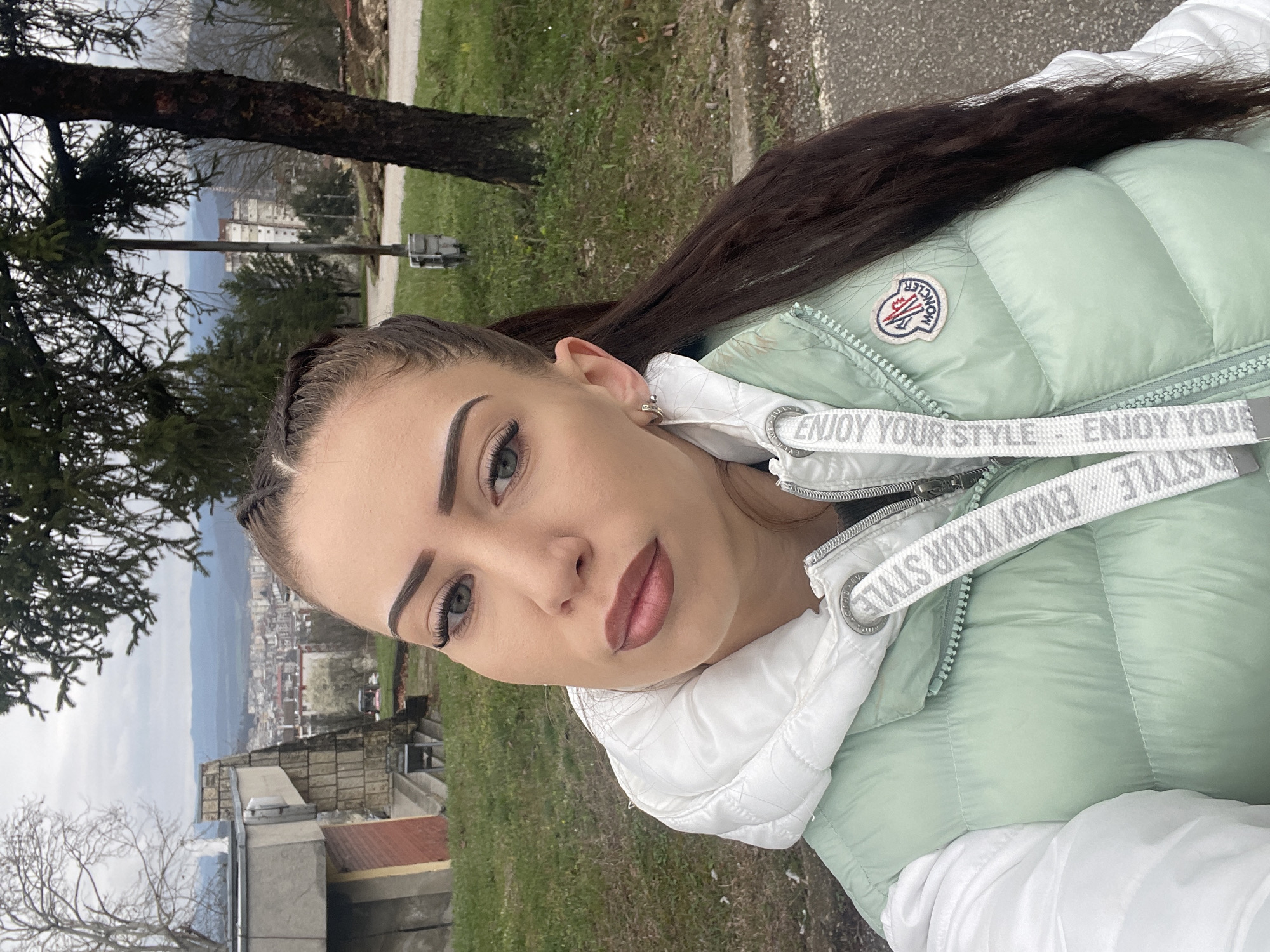 A young woman is photographed at eye level in an outdoor setting. Her head and shoulders are prominently featured in the foreground. She has long, dark hair and is wearing a light green puffy jacket with white accents. A white strap with the text "ENJOY YOUR STYLE" is visible across her chest. In the background, the urban landscape of Veliko Tarnovo, Bulgaria, is discernable. A building with multiple stories and a mix of overcast sky is visible. Trees are also present in the background, adding to the overall natural and urban environment. The image is slightly rotated, which gives it a casual and candid feel.
