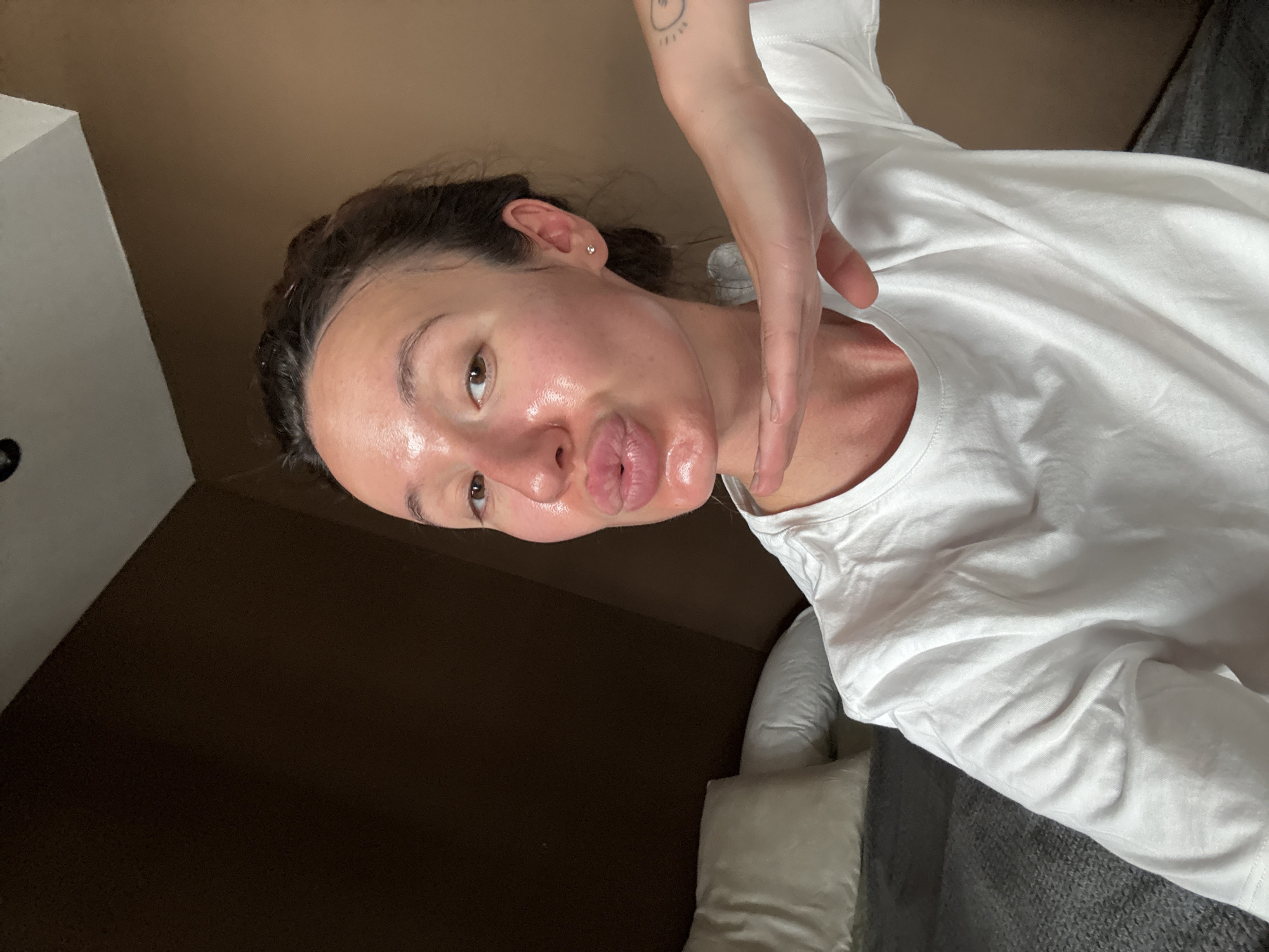 An adult female is depicted in a medium shot, posing indoors. She has dark hair pulled back and brown eyes. Her face and lips are coated in a clear, highly reflective substance, giving her skin a glossy appearance. She is puckering her lips in a "kissing face" expression. She wears a plain white crew-neck t-shirt. A small silver stud earring is visible in her right ear. Her left hand is positioned horizontally under her chin, palm facing upwards. A small, circular tattoo with radiating lines is visible on her left forearm. In the background, a bed is visible with at least two white pillows and a grey textured blanket or duvet. The room features walls painted in varying shades of brown, with a white, angled ceiling visible at the top. A dark circular object is affixed to the white ceiling. The scene is located in Tel-Aviv, Israel.